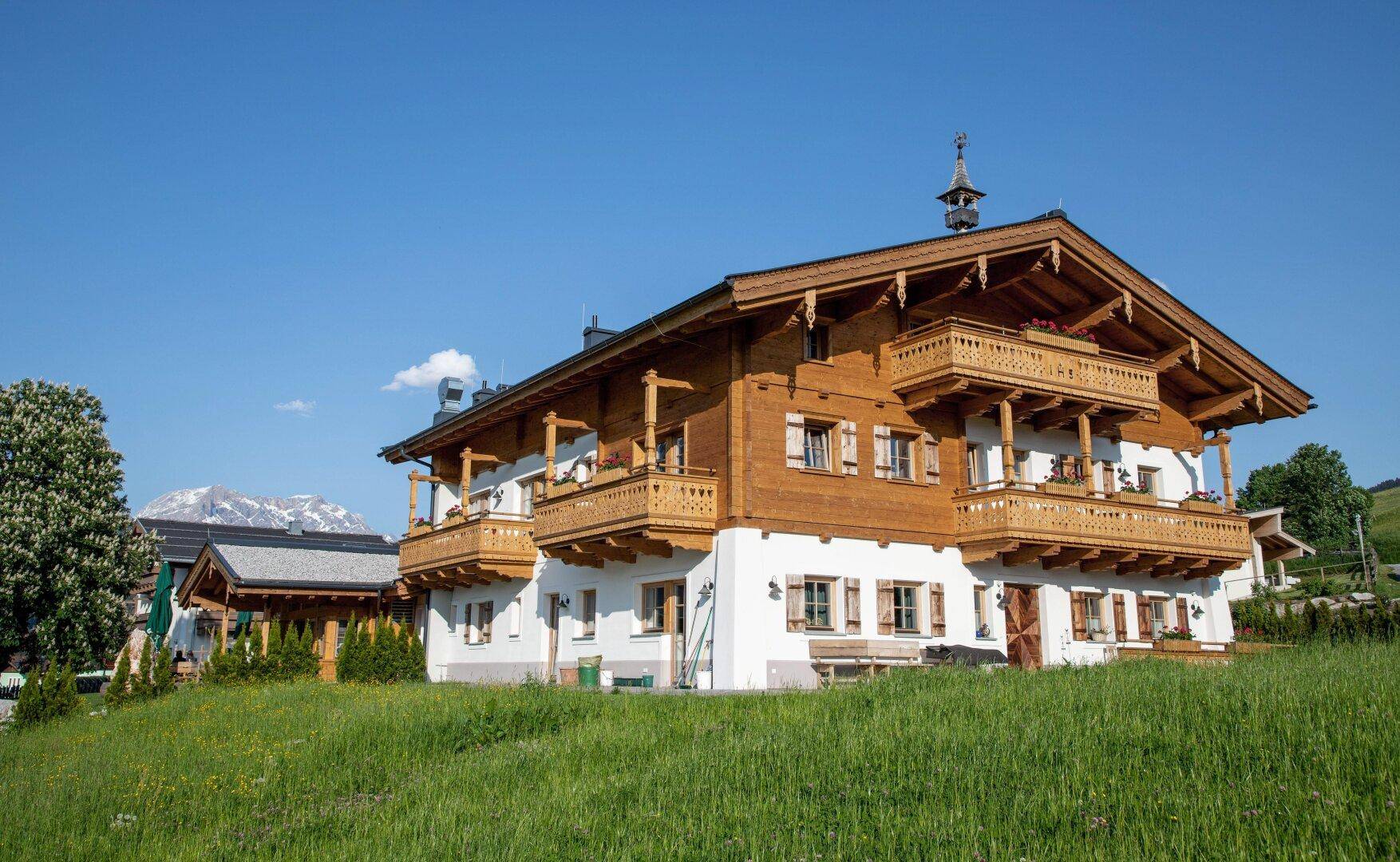Traditionelles Alpenhaus mit Holzbalkonen, verzierten Geländern und weißer Fassade vor grüner Landschaft.