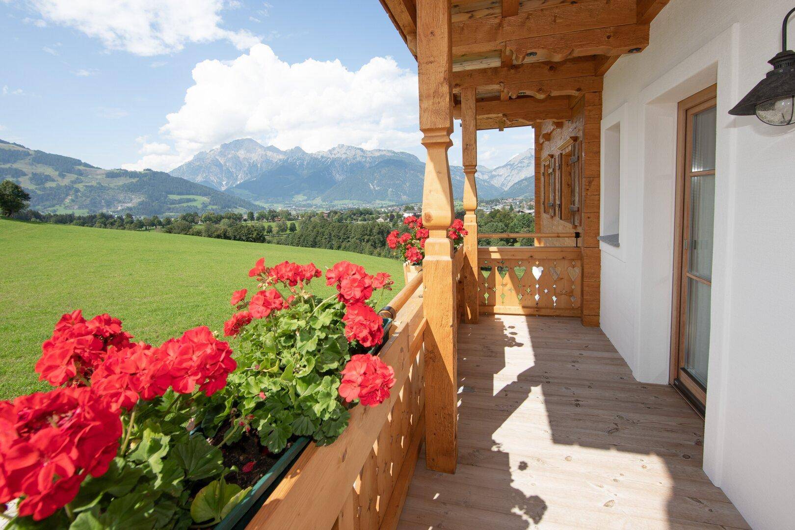 Holzbalkon mit roten Geranien und weitem Blick über grüne Hügel und Berge.