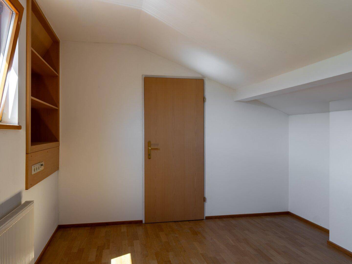 Helles Zimmer im Dachgeschoss mit Holzboden, Dachschräge und Fenster.