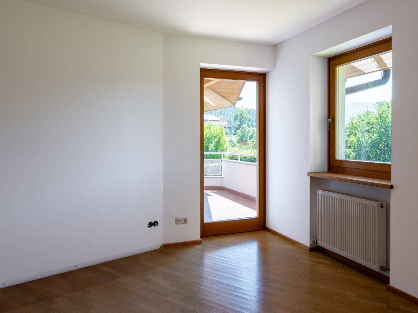 Schlafzimmer mit Holzboden, Heizkörper und Zugang zu einem sonnigen Balkon.