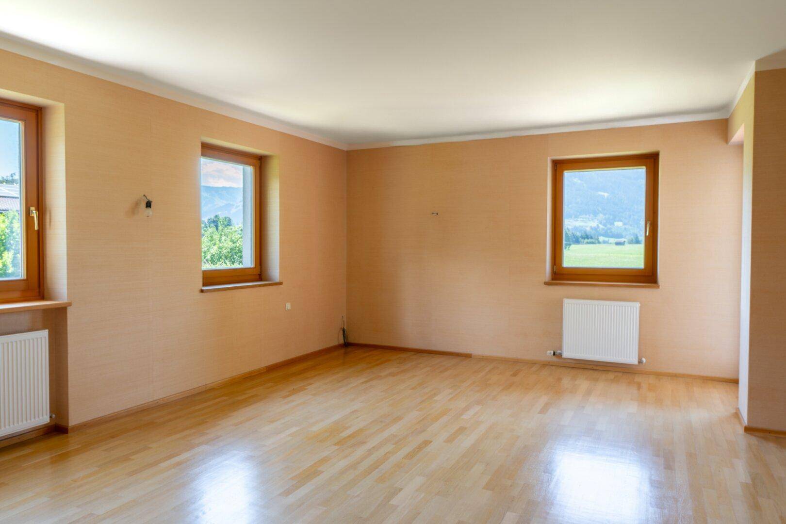 Geräumiges Zimmer mit Holzboden, hellen Wänden und Fenstern mit Bergblick.