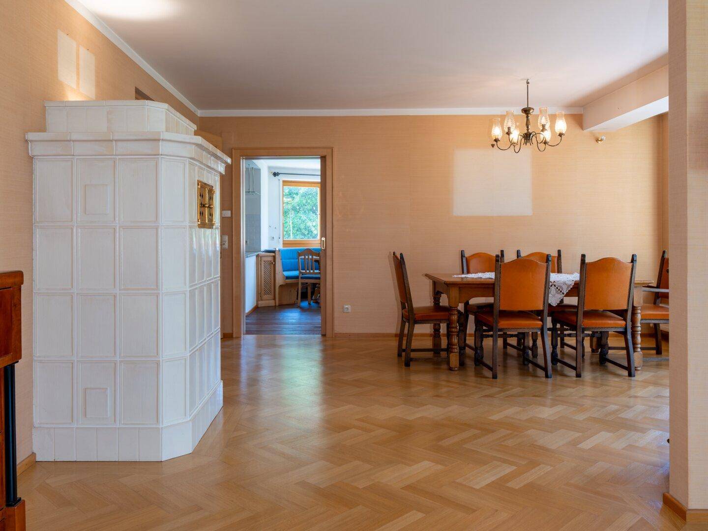 Esszimmer mit traditionellem Kachelofen, Holzboden und Blick in die angrenzende Küche.