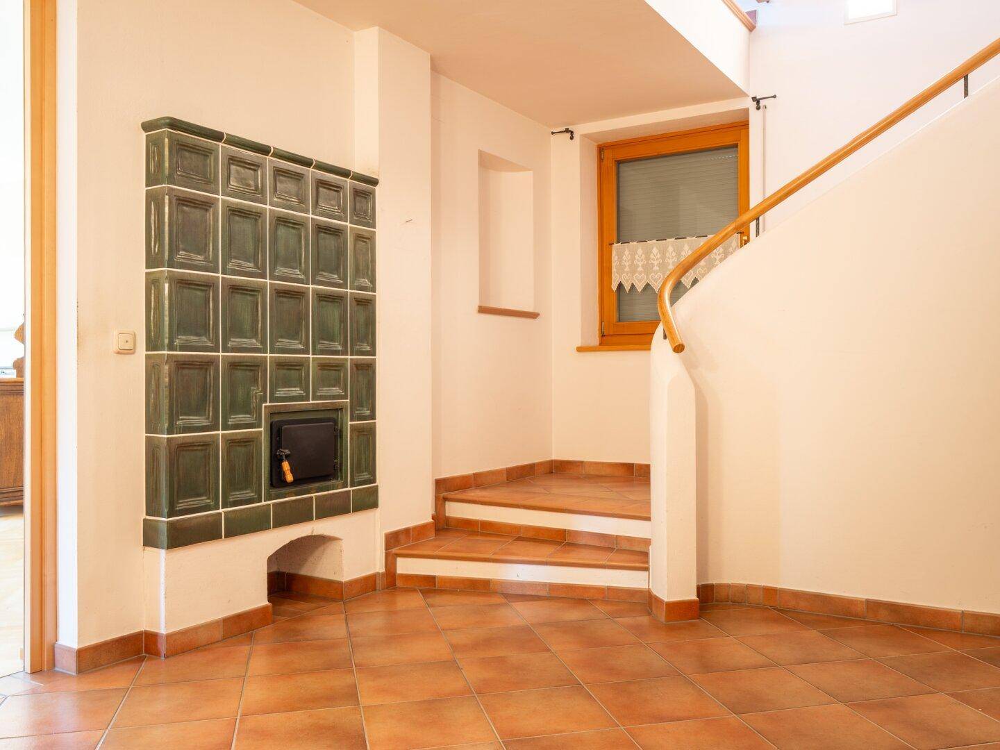 Heller Flur mit Terrakottafliesen, Holztreppe und einem grünen Kachelofen.