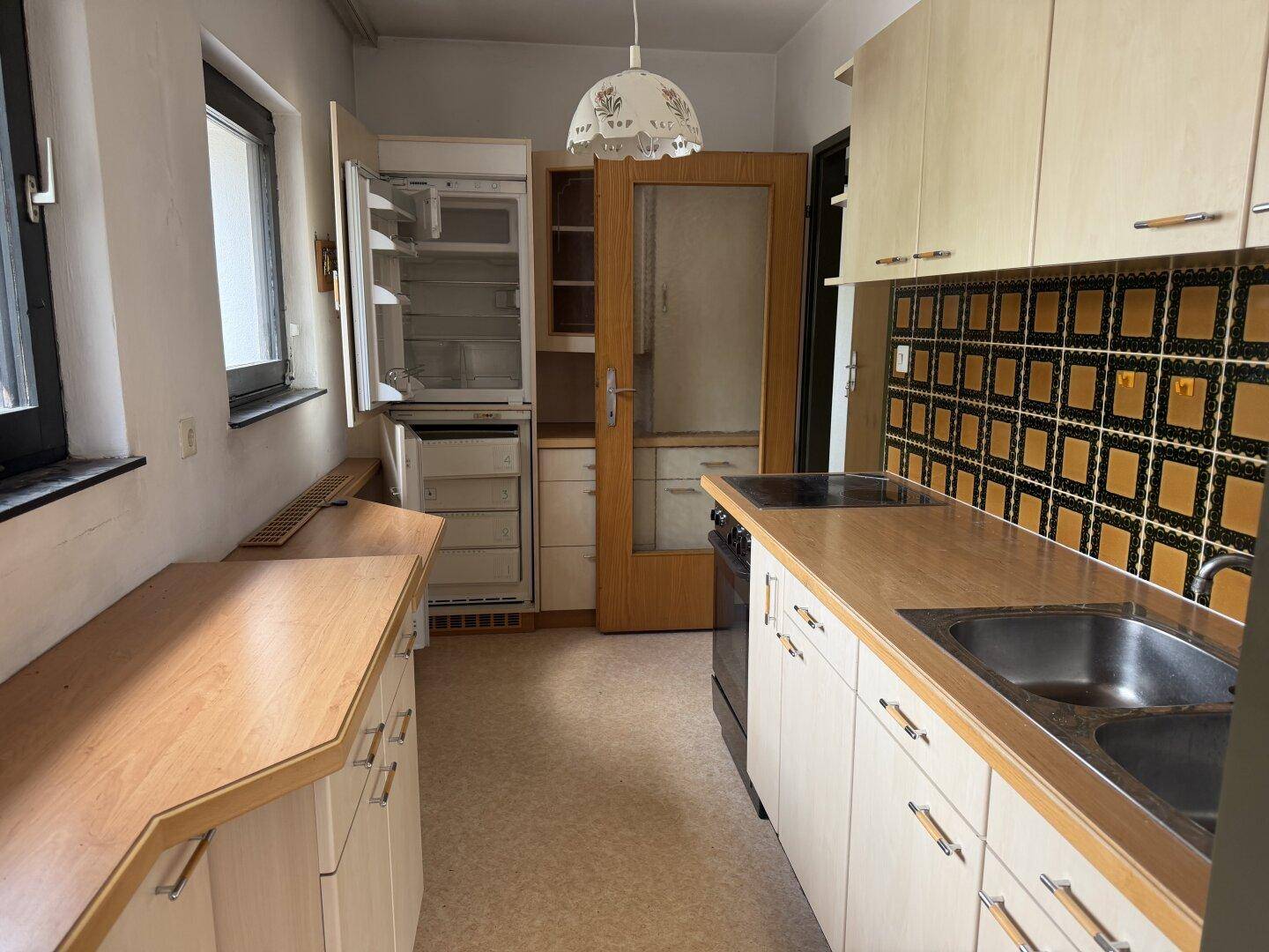 Funktionale Küche mit hellen Holzschränken, integriertem Kühlschrank und charakteristischen Wandfliesen.