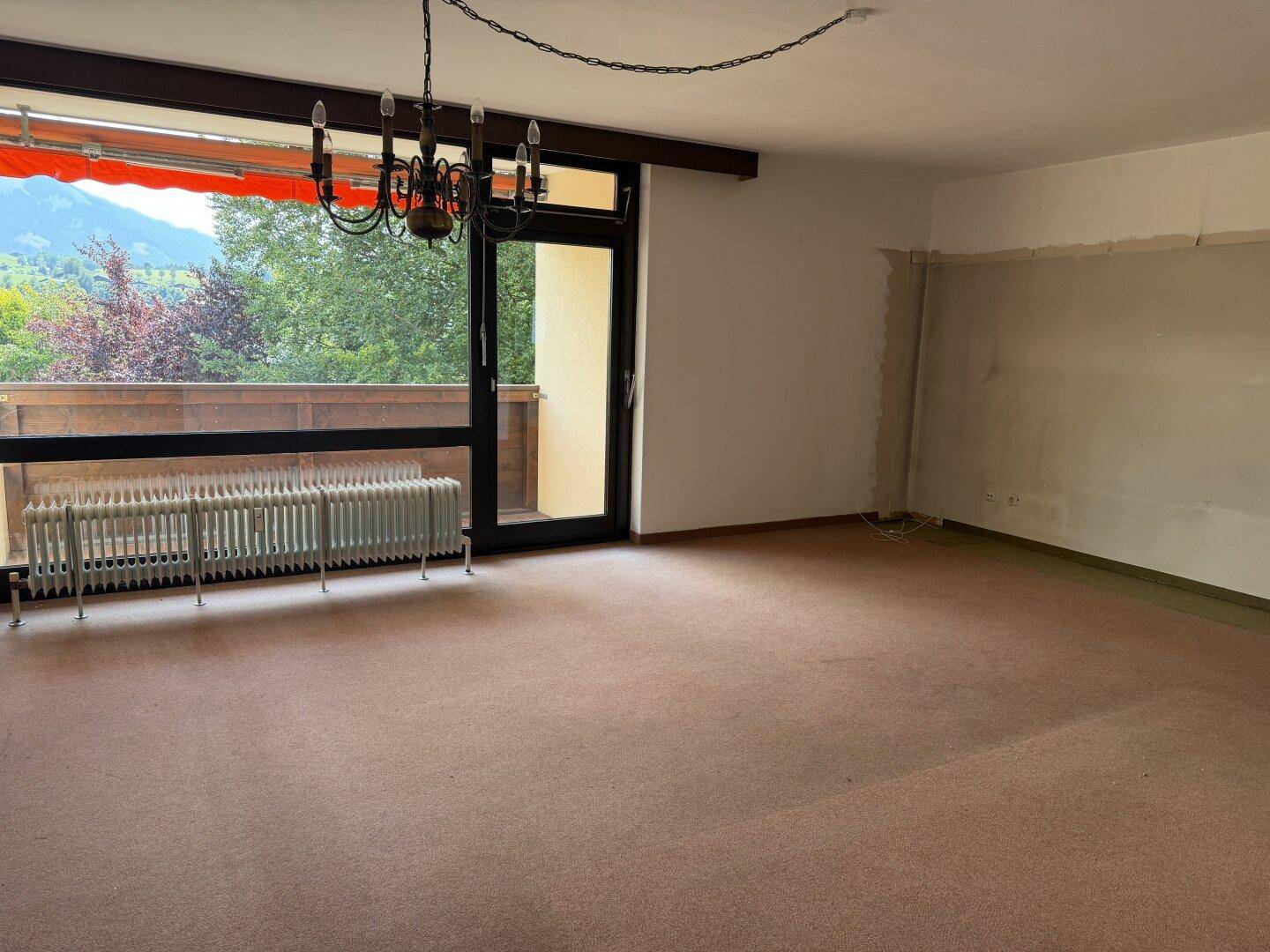 Geräumiges Wohnzimmer mit Teppichboden, großem Fenster und Zugang zum Balkon mit Bergblick.