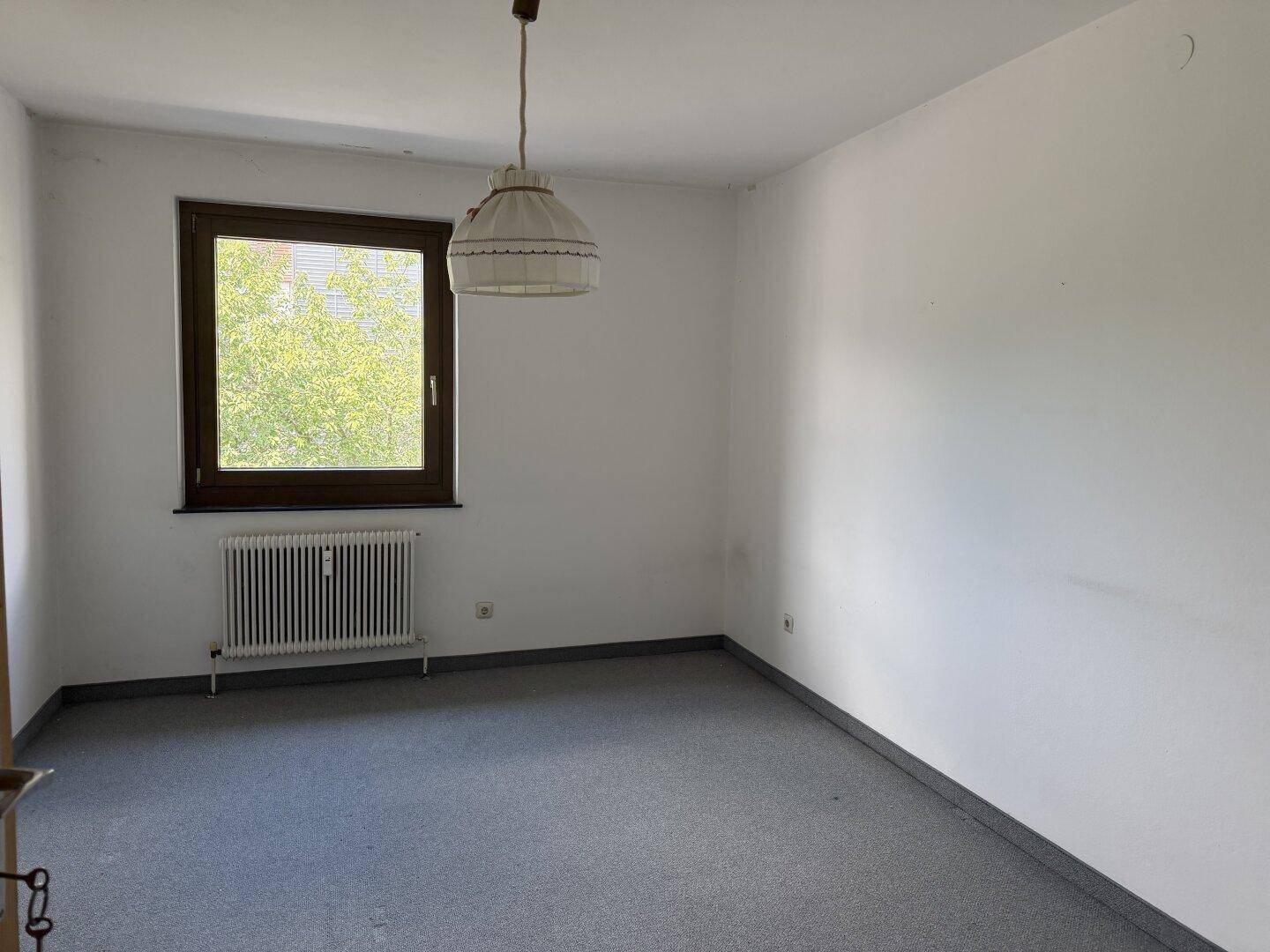 Freundliches Zimmer mit grauem Teppichboden, Fenster und Heizkörper, geeignet als Schlaf- oder Gästezimmer.