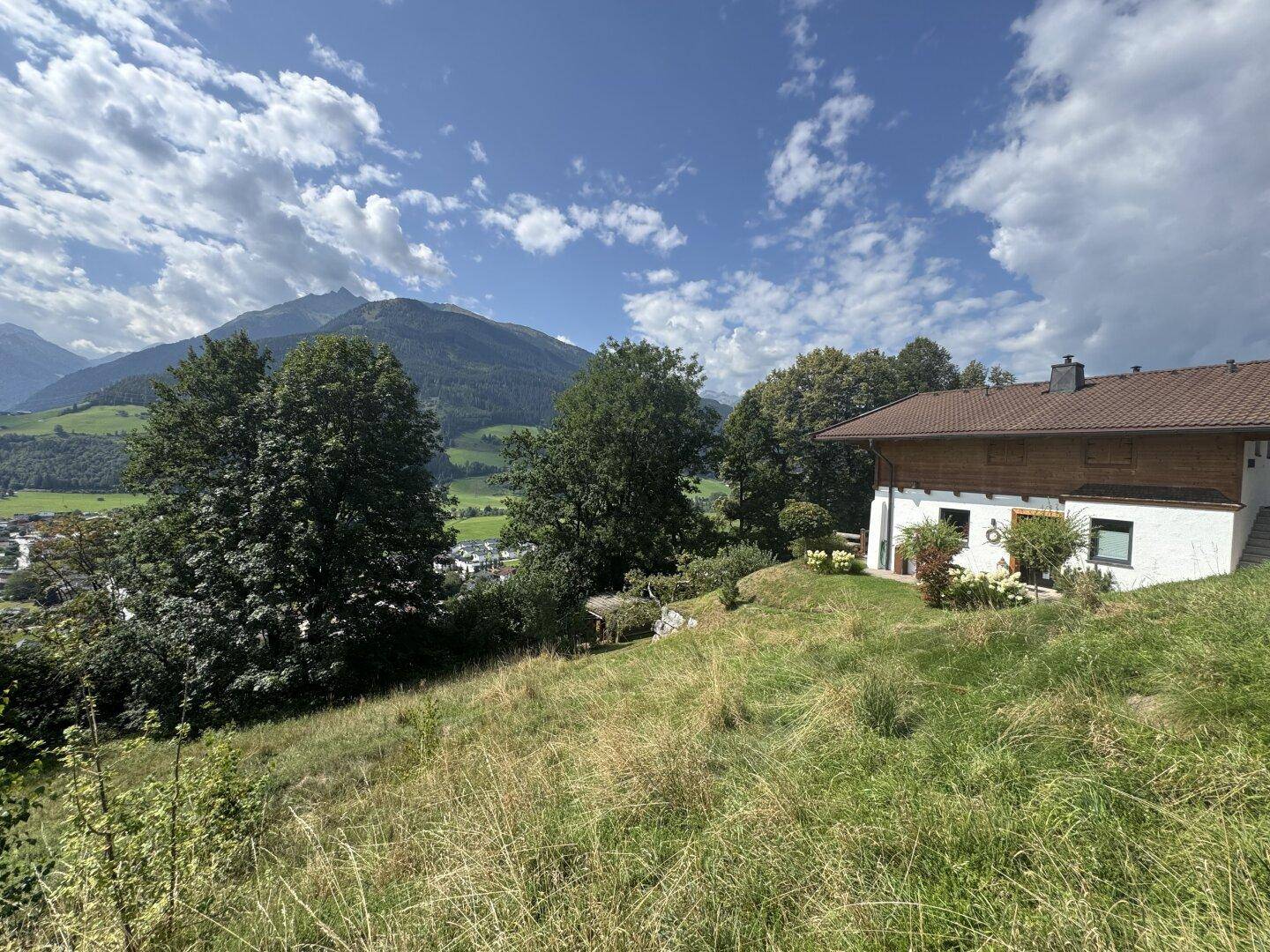 Charmantes Haus mit weißer Fassade und Holzverkleidung, eingebettet in eine grüne Hügellandschaft mit Bergblick.