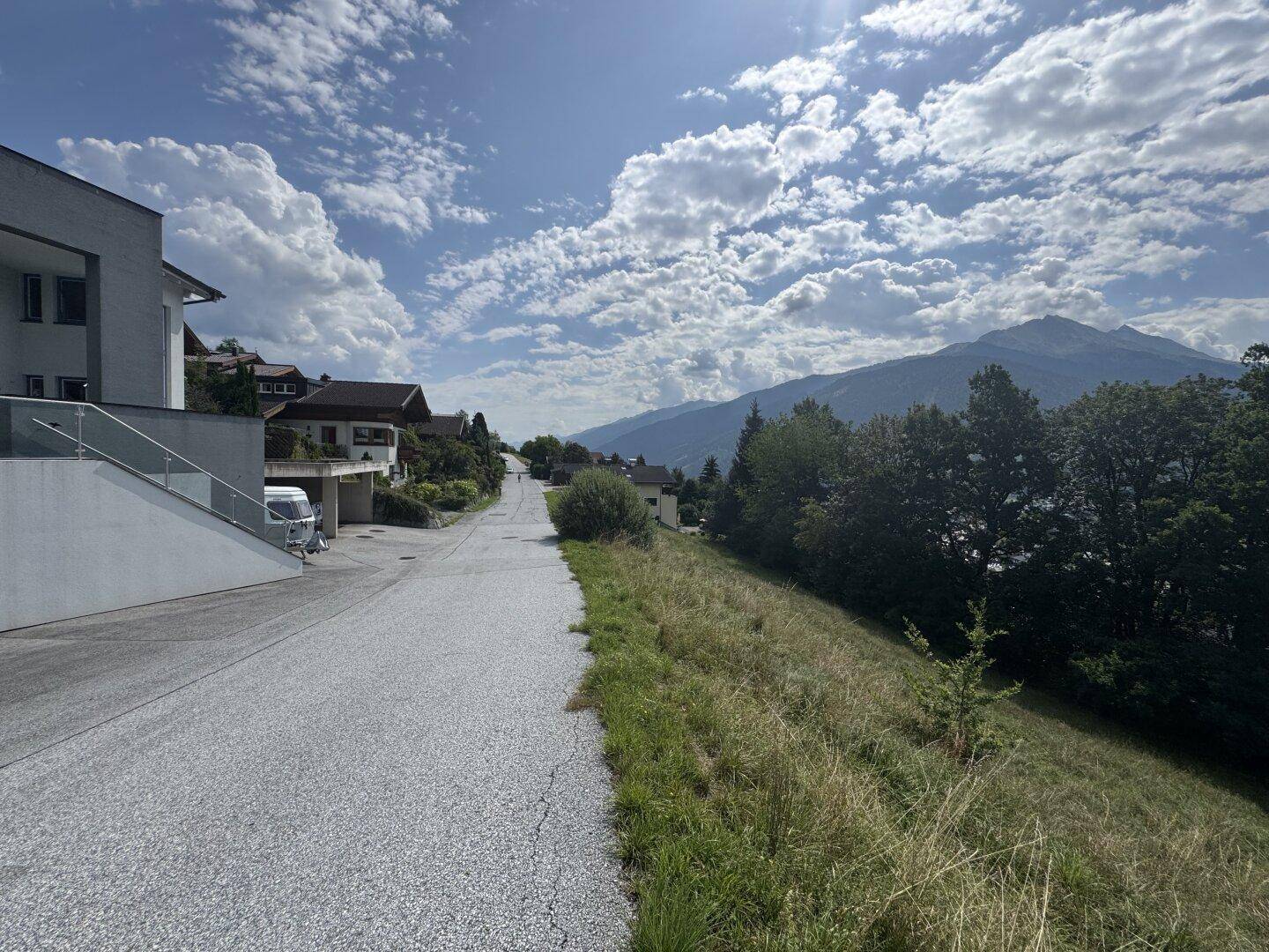 Eine ruhige Straße führt bergauf, gesäumt von Häusern und einer grünen Landschaft mit Bergblick.