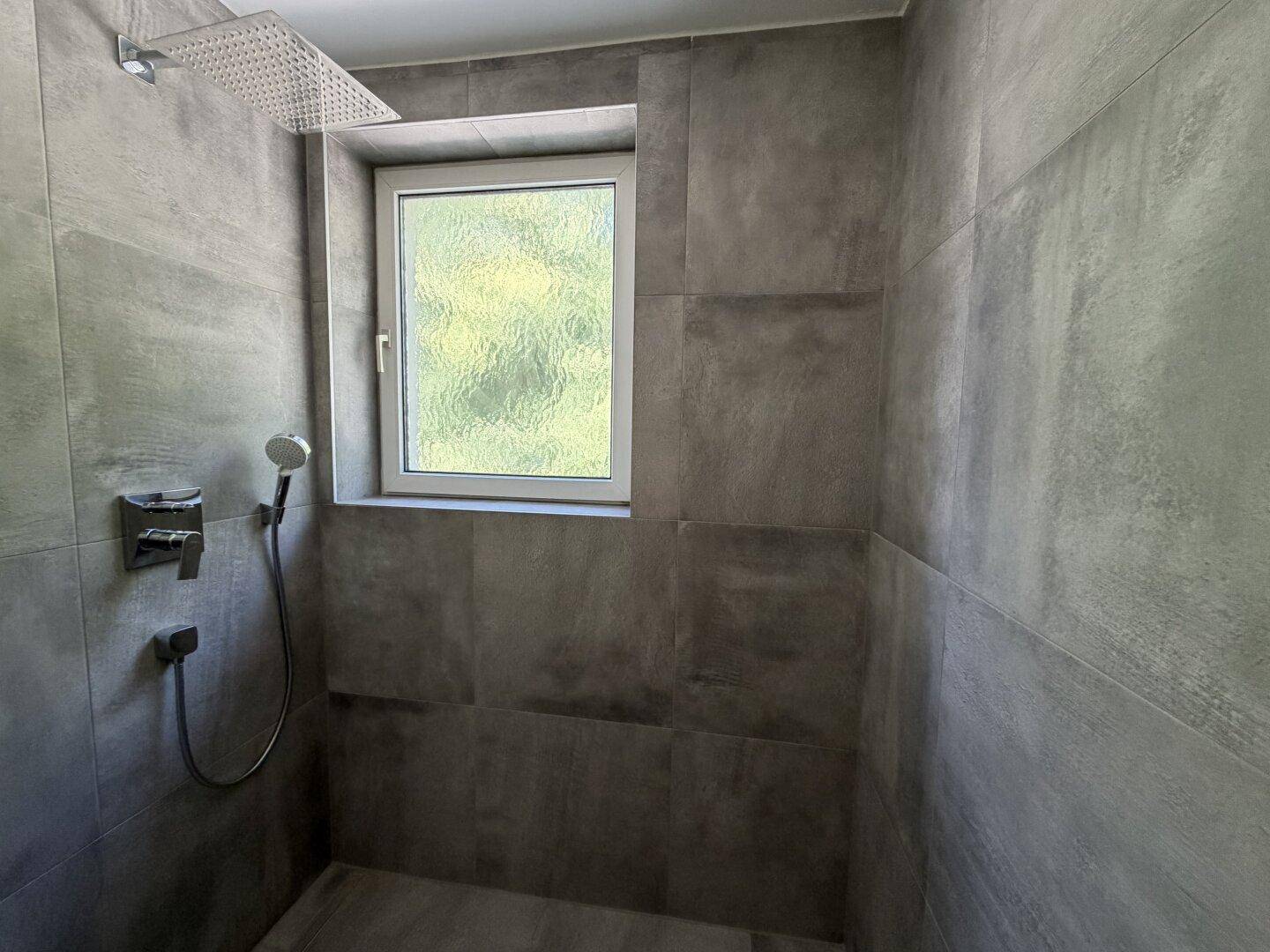 Moderne Duschkabine mit grauen Fliesen, Regendusche und einem Fenster für Tageslicht.