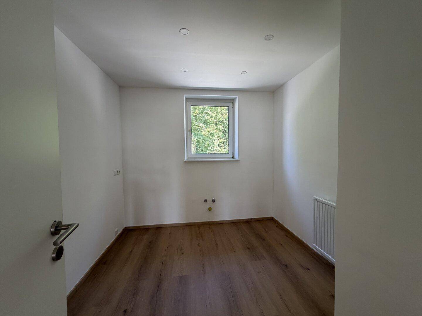 Helles, leeres Zimmer mit Holzboden und Fenster, ideal als Schlaf- oder Arbeitszimmer.
