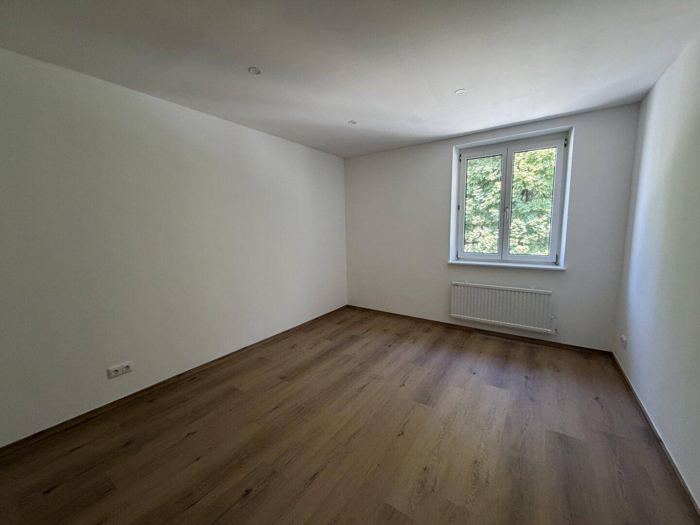 Geräumiges, leeres Schlafzimmer mit Holzboden und großem Fenster für viel Tageslicht.