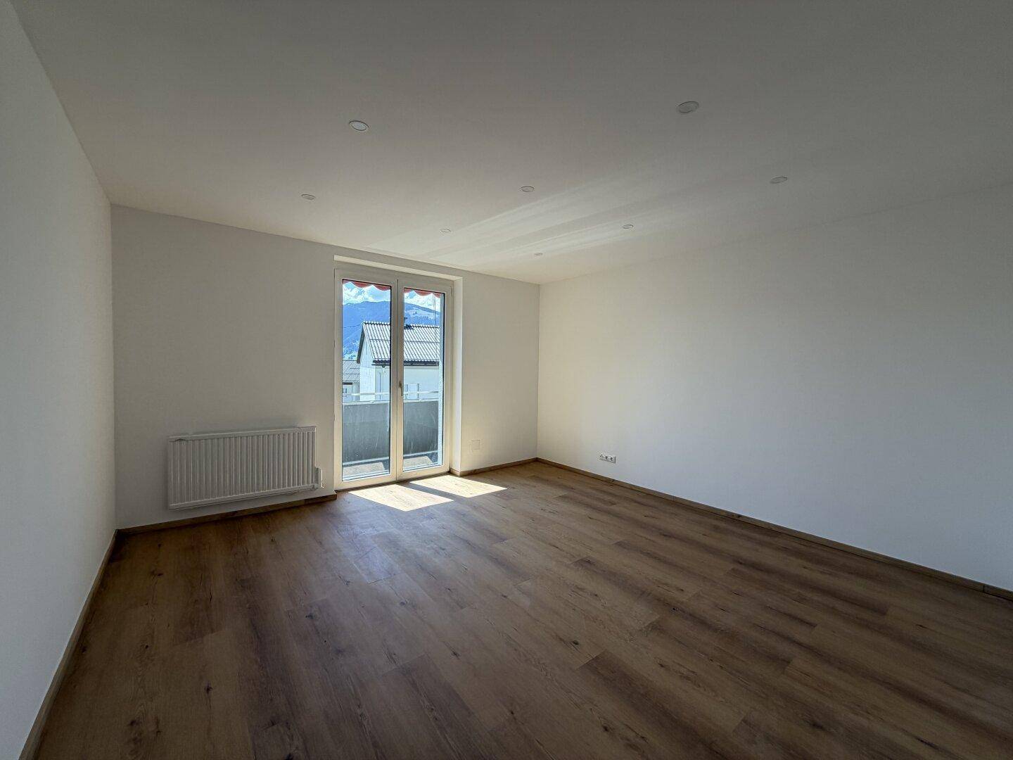 Helles, leeres Wohnzimmer mit Holzboden und Zugang zu einem Balkon oder einer Terrasse.