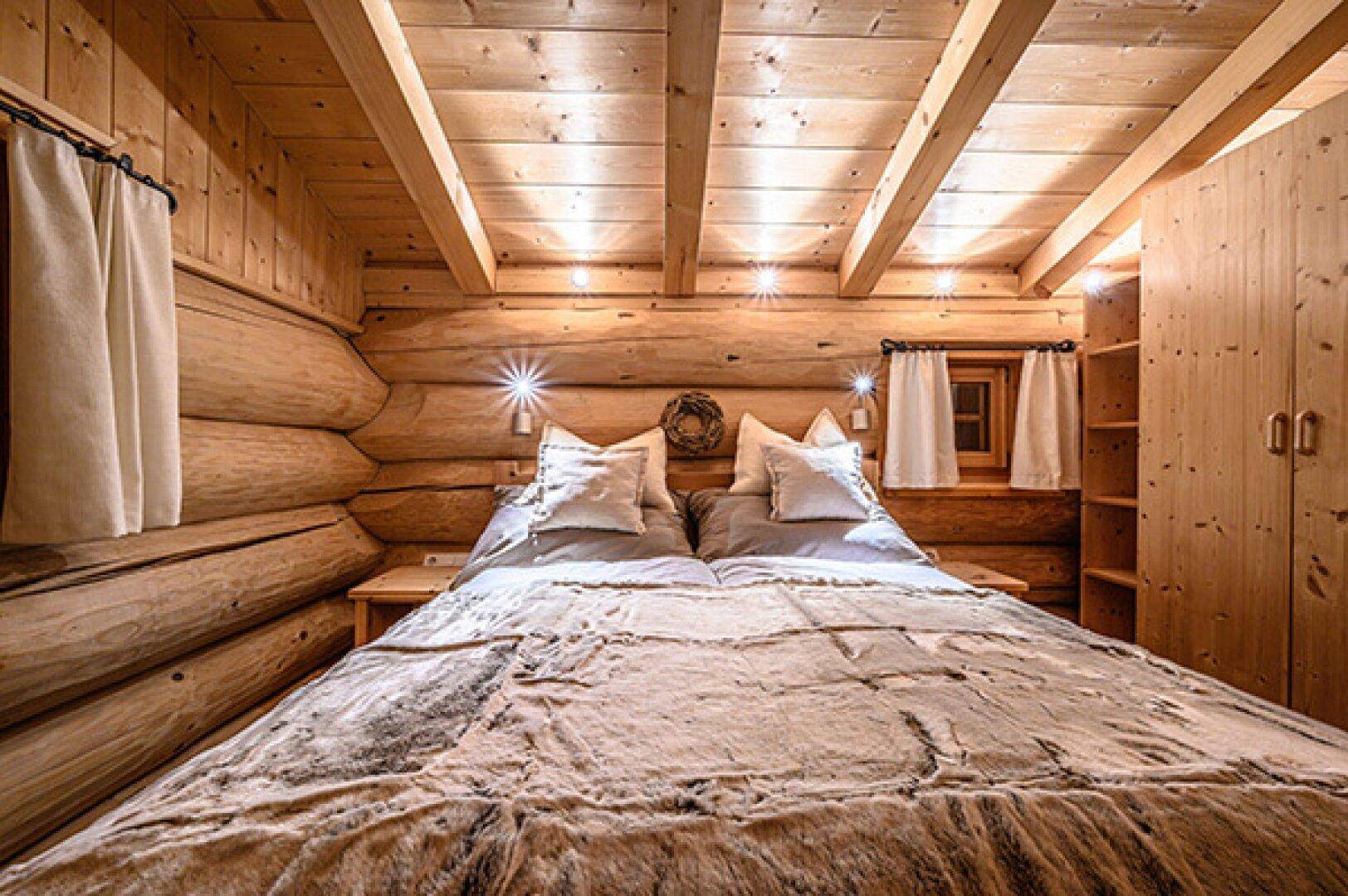 Schlafzimmer in einem Holzhaus mit Doppelbett, Holzbalkendecke und indirekter Beleuchtung.