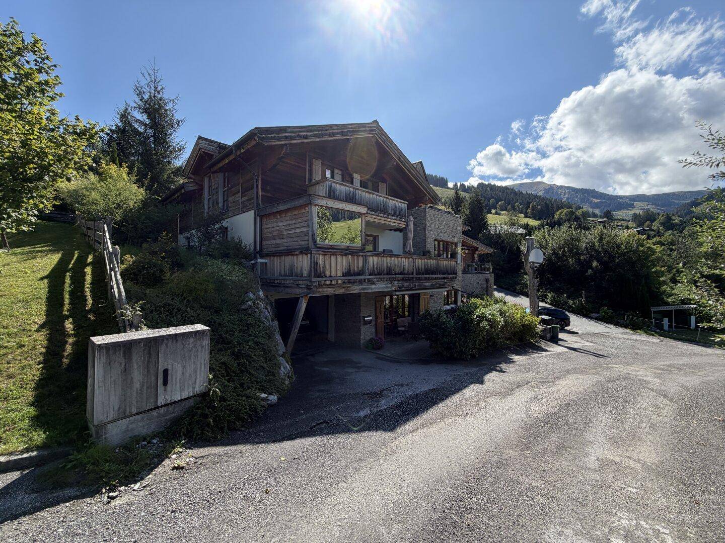 Gebäude mit Holz- und Steinfassade, Balkonen und einer Zufahrt in bergiger Landschaft.
