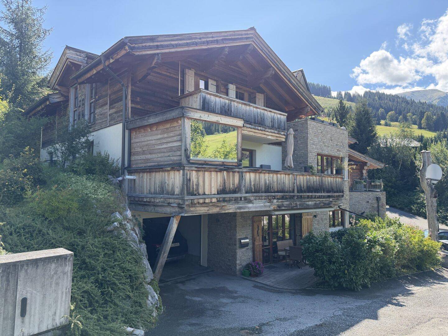 Gebäude mit Holz- und Steinfassade, Balkonen und einer Zufahrt in sonniger Umgebung.