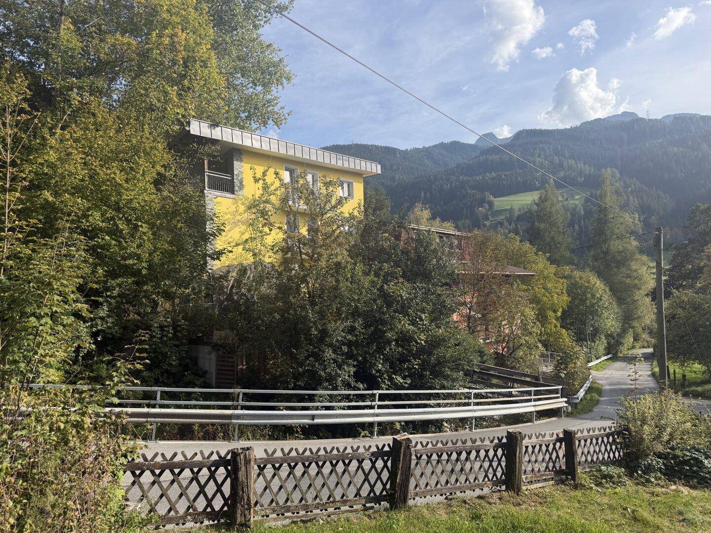 Gebäudekomplex eingebettet in eine grüne Berglandschaft mit Bäumen und einer kurvigen Straße im Vordergrund.