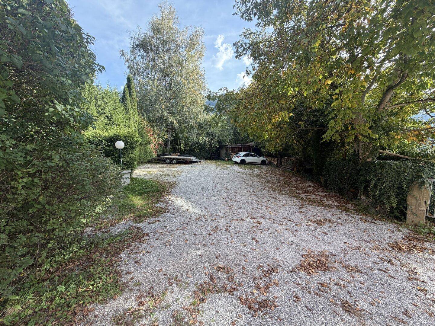 Großer Schotterparkplatz umgeben von Bäumen und Sträuchern mit zwei geparkten Autos und einem Anhänger.