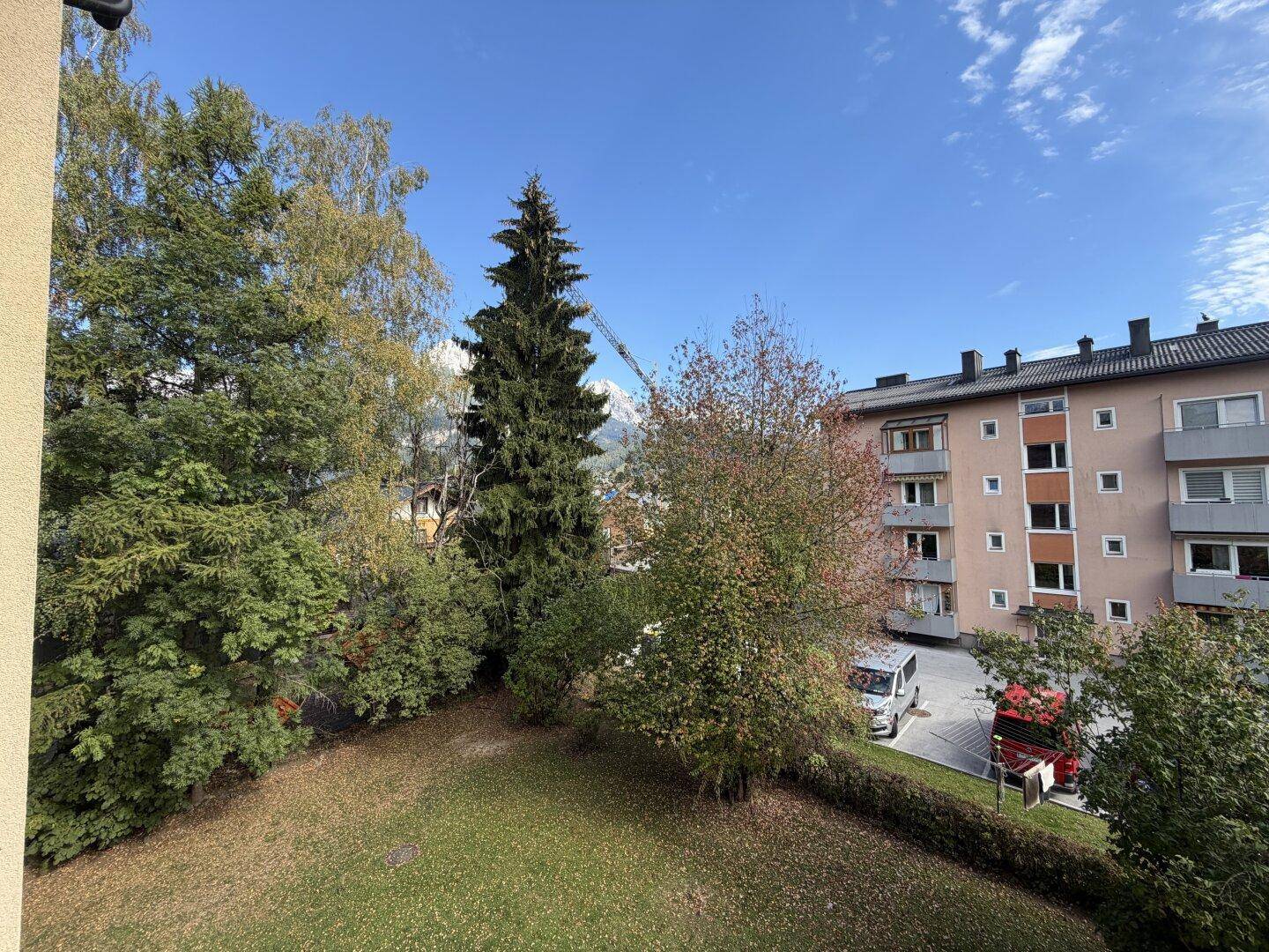 Weitläufiger Blick ins Grüne mit Bäumen, einem Wohngebäude und Bergen im Hintergrund unter blauem Himmel.