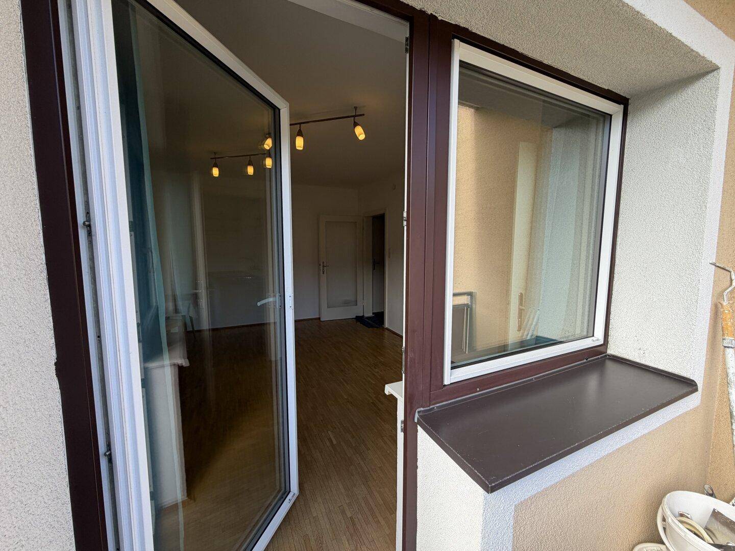 Blick vom Innenraum auf einen Balkon mit braunen Fensterrahmen und einer geöffneten Balkontür.