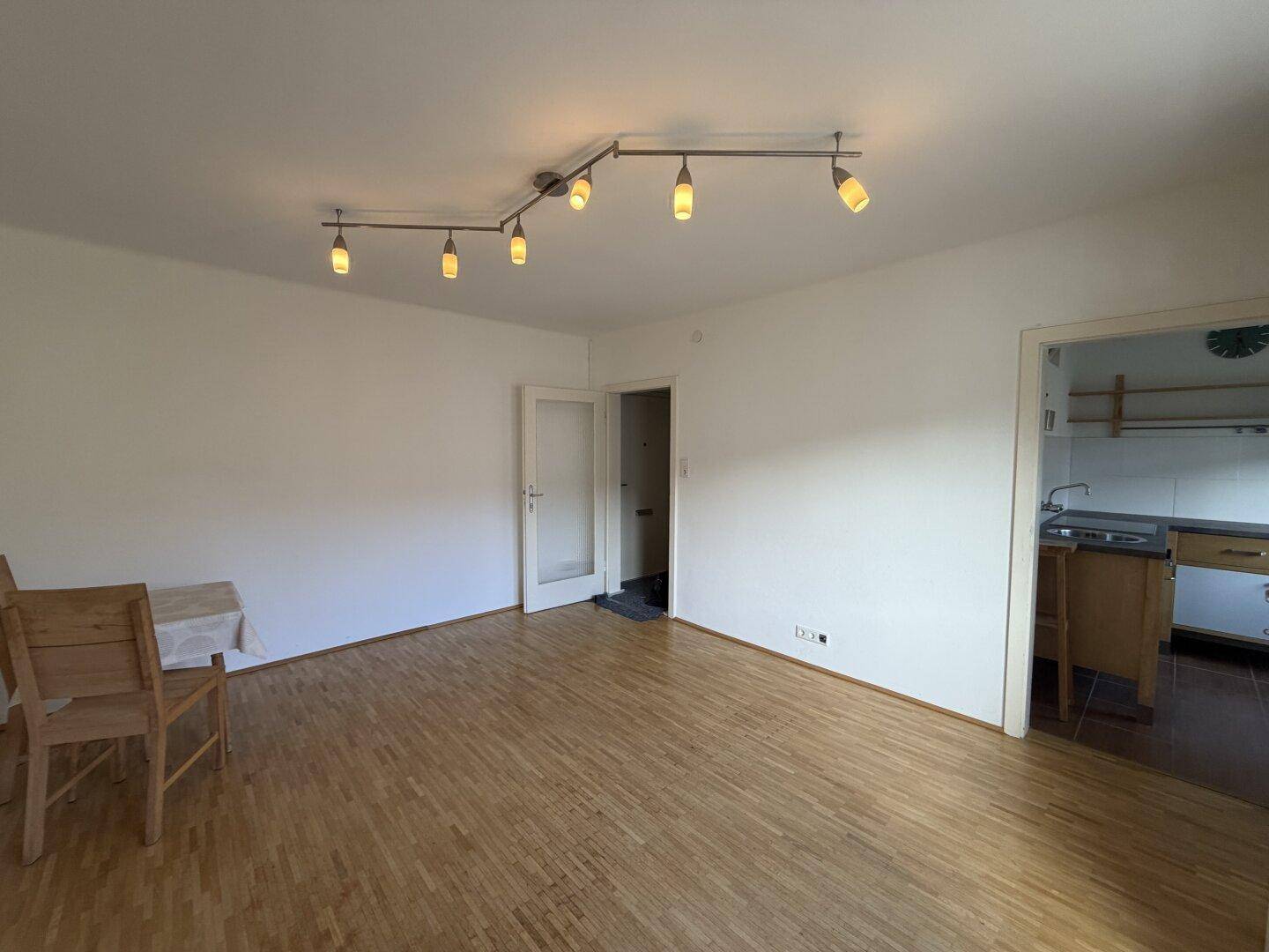 Geräumiges Wohnzimmer mit hellem Parkettboden und direktem Zugang zur Küche durch eine offene Tür.