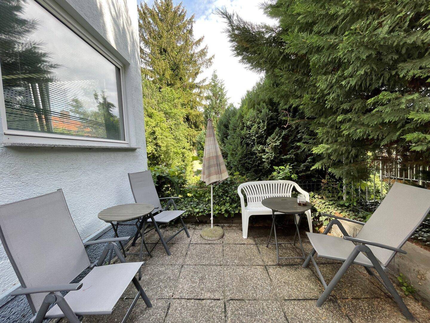 Gemütliche gepflasterte Terrasse mit Gartenmöbeln und Sonnenschirm, umgeben von Bäumen.