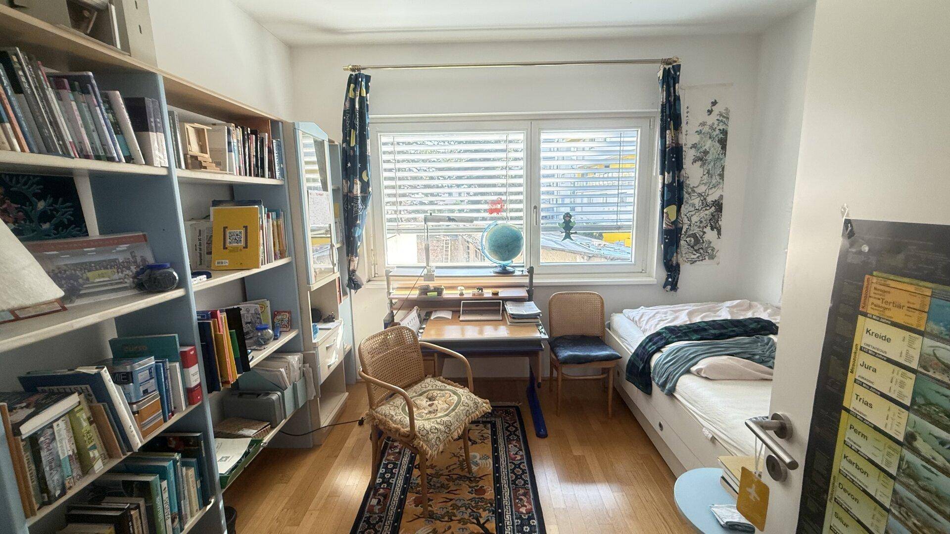 Helles Schlafzimmer mit Holzboden, Bücherregal und Schreibtisch am Fenster.