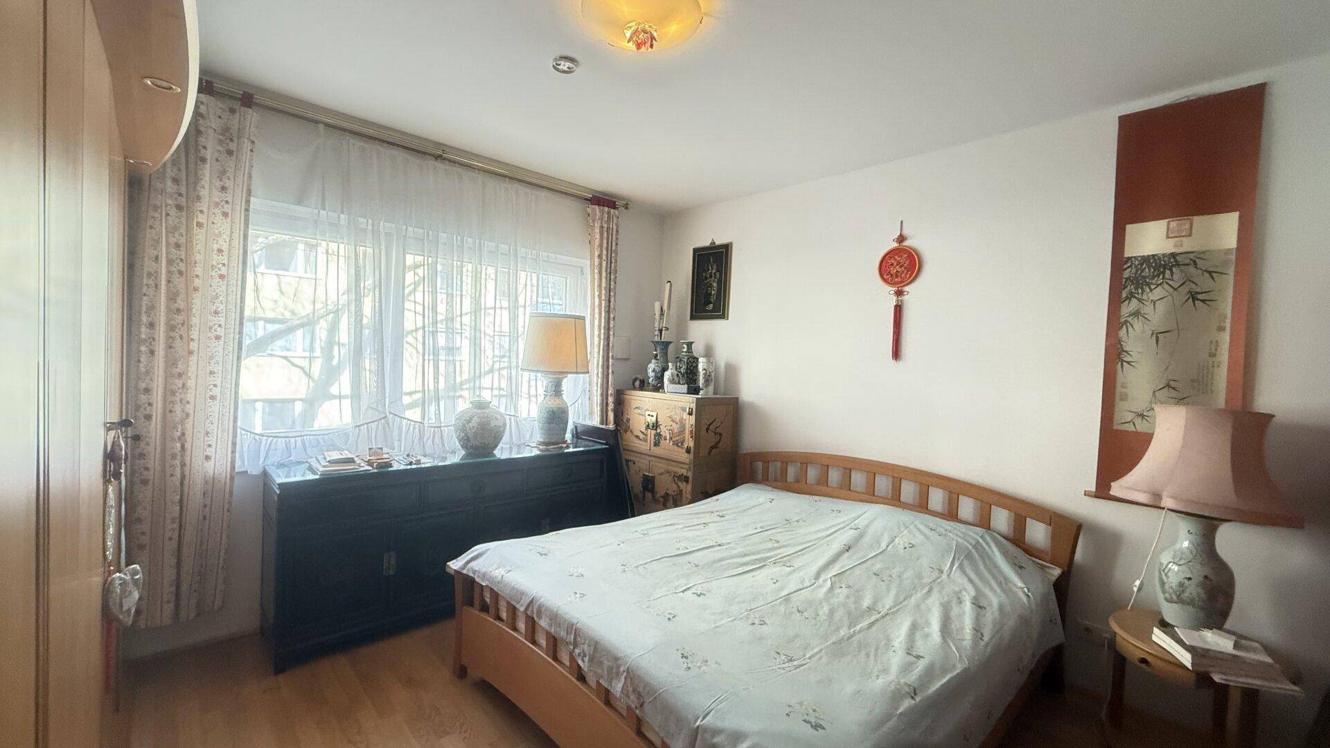 Schlafzimmer mit Doppelbett, Kommode und Fenster mit Vorhängen.