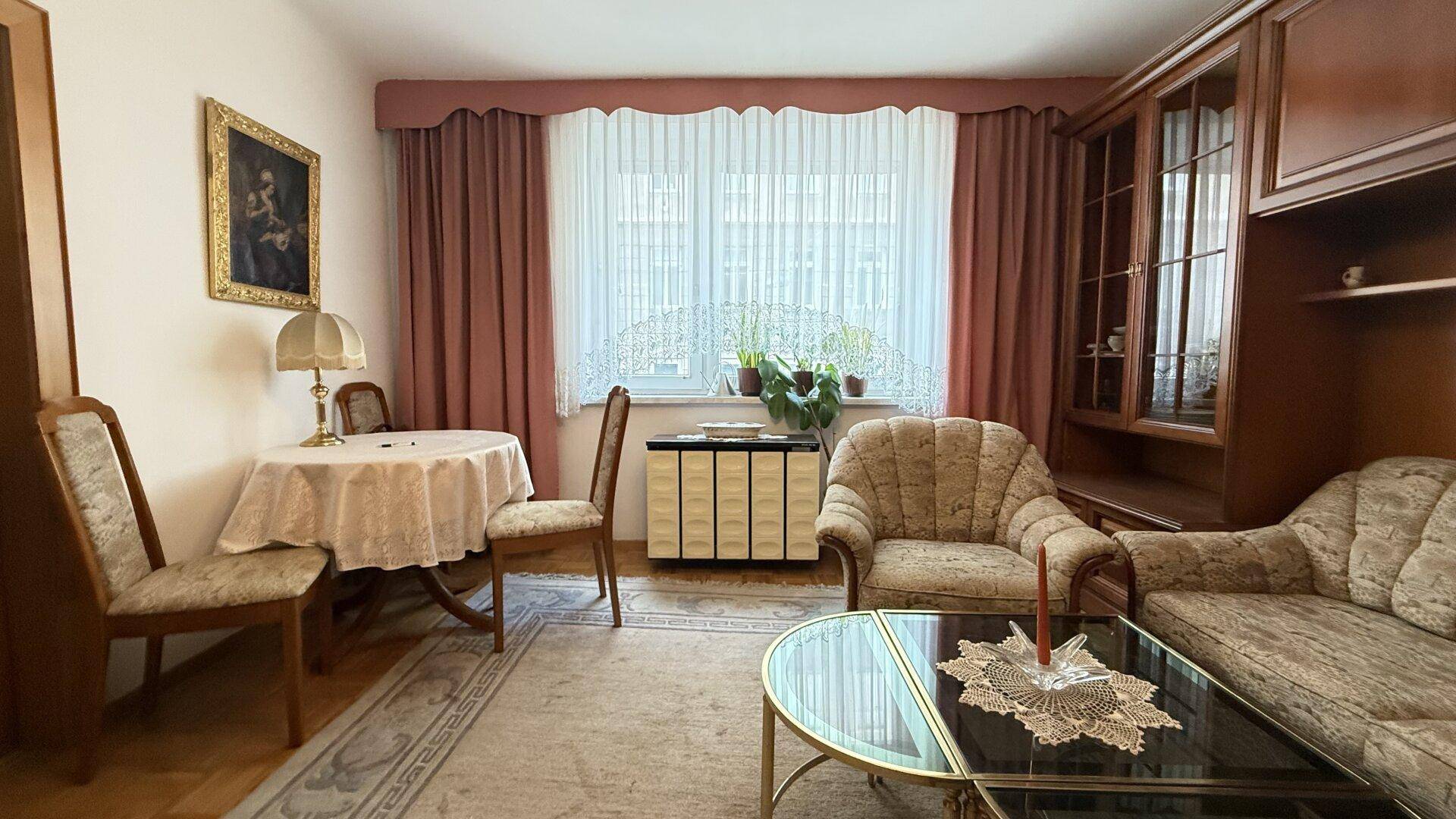 Gemütliches Wohnzimmer mit traditionellen Möbeln, Teppichboden und einem großen Fenster mit Vorhängen.
