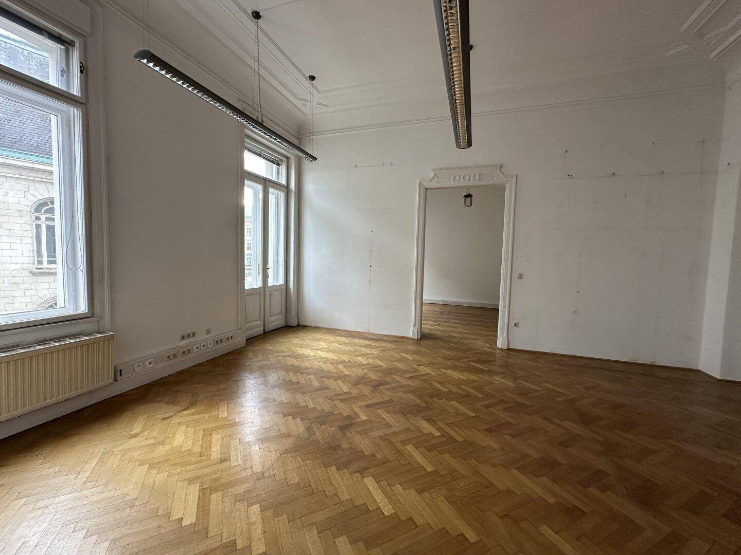 Heller Raum mit Fischgrätparkett, hohen Fenstern und dekorativen Stuckelementen an der Decke.