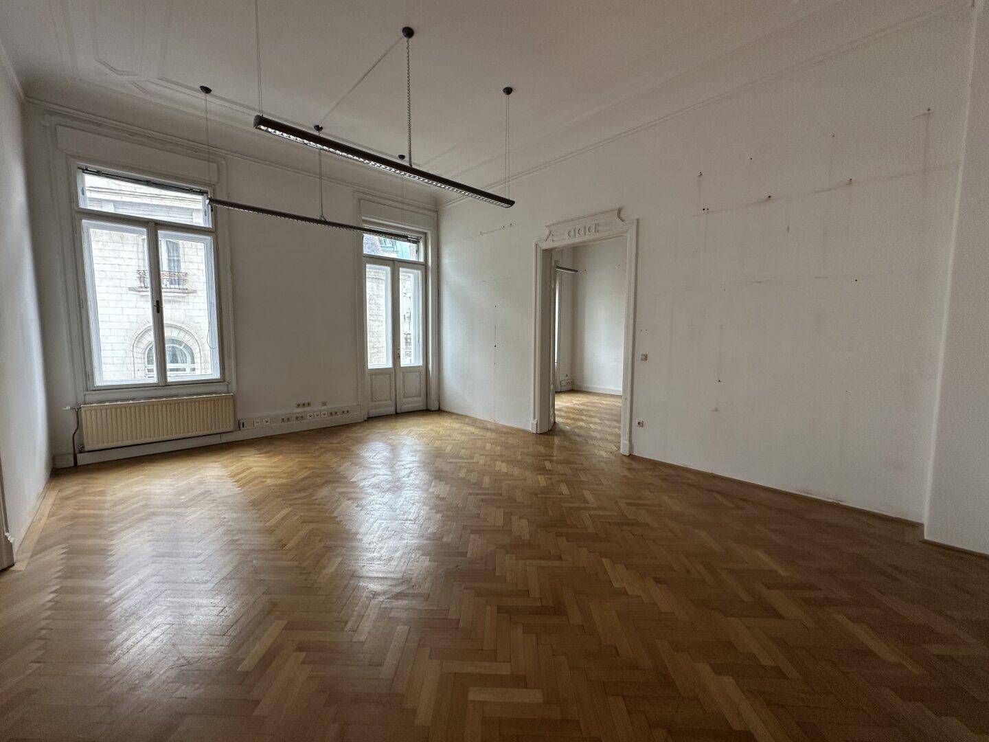 Heller Raum mit klassischem Fischgrätparkett, hohen Fenstern und einer breiten Türöffnung zu einem weiteren Zimmer.