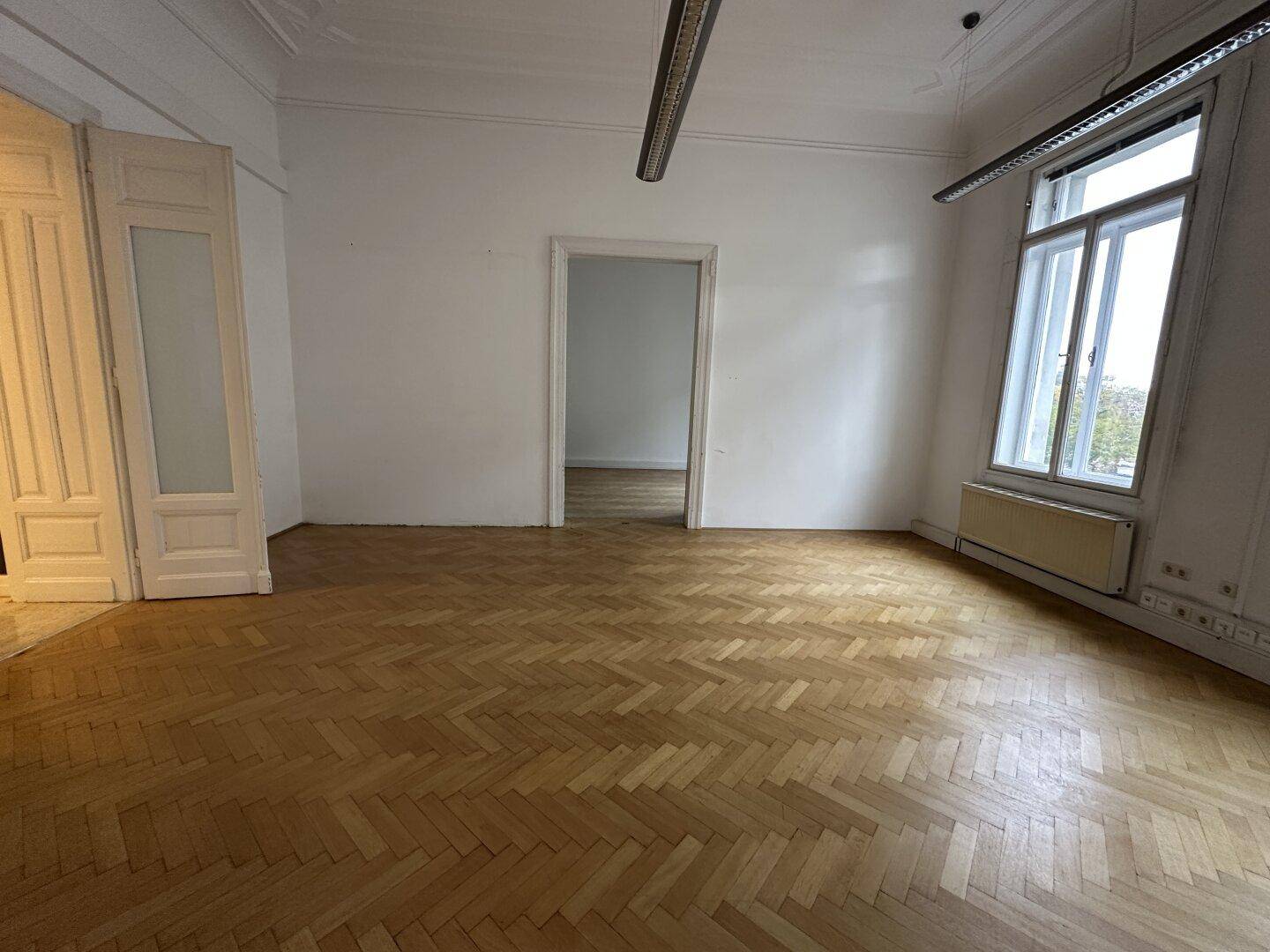 Heller Raum mit Fischgrätparkett, hohen Fenstern und einer Türöffnung zu einem angrenzenden Zimmer.