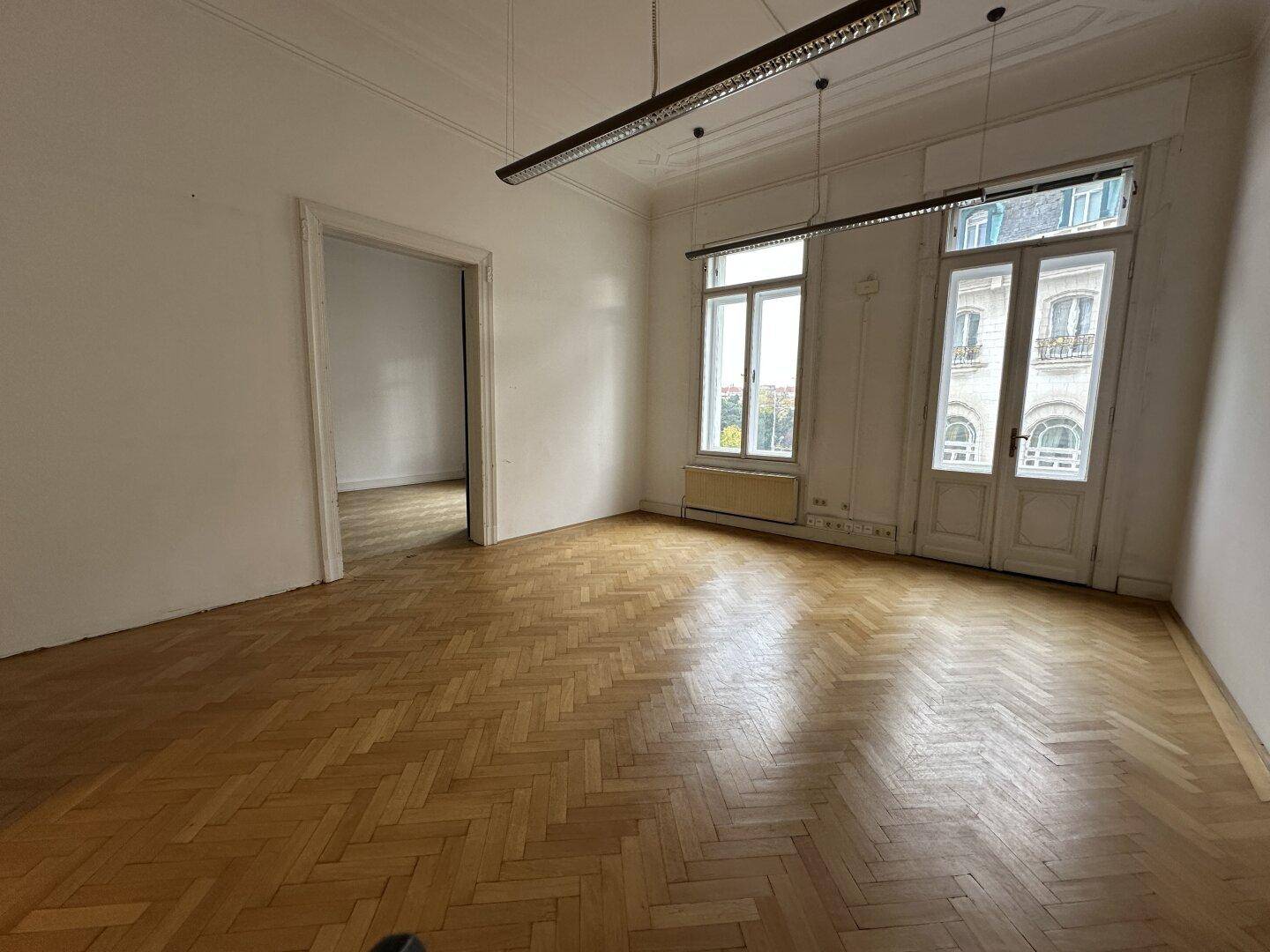 Geräumiger Raum mit Fischgrätparkett, hohen Fenstern und einer breiten Türöffnung zu einem weiteren Zimmer.