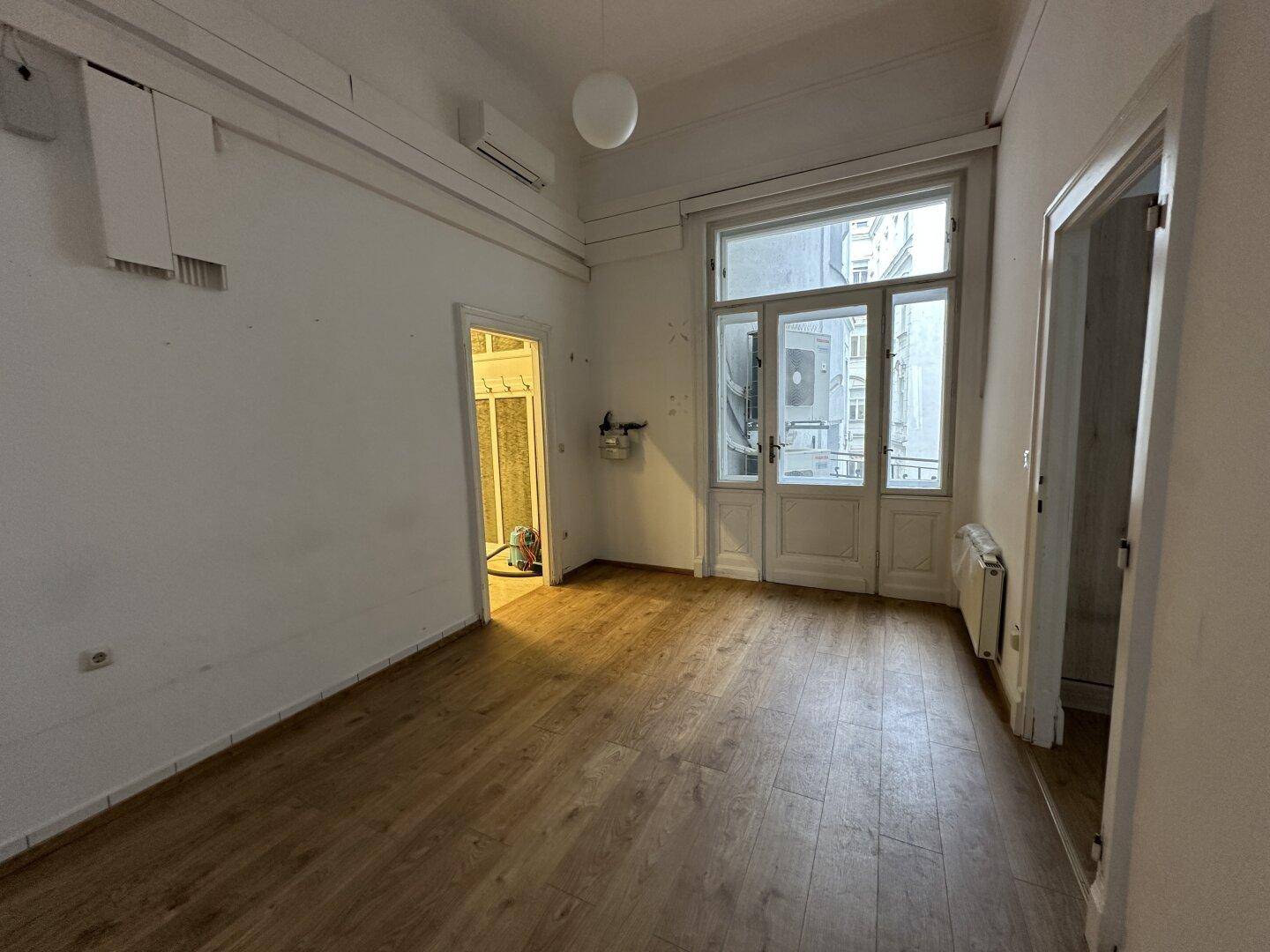 Heller Raum mit Laminatboden, hohen Fenstern und einer Klimaanlage an der Decke.