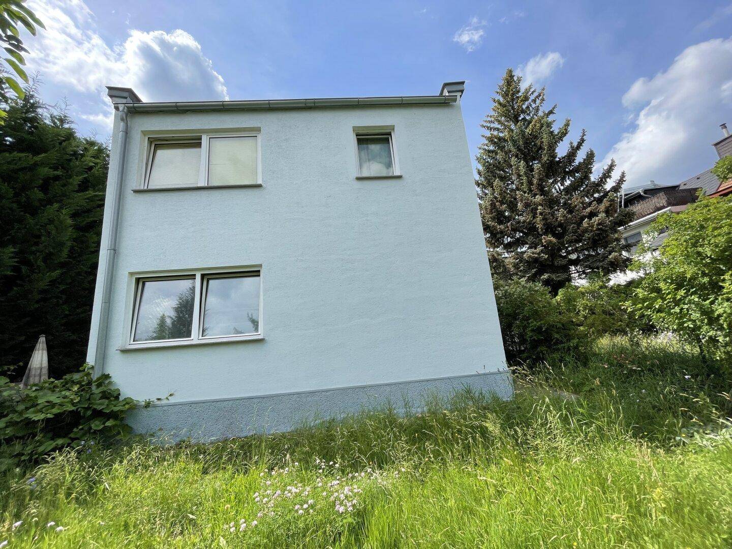 Rückansicht des Gebäudes mit hellblauer Fassade und Fenstern, umgeben von viel Grün.