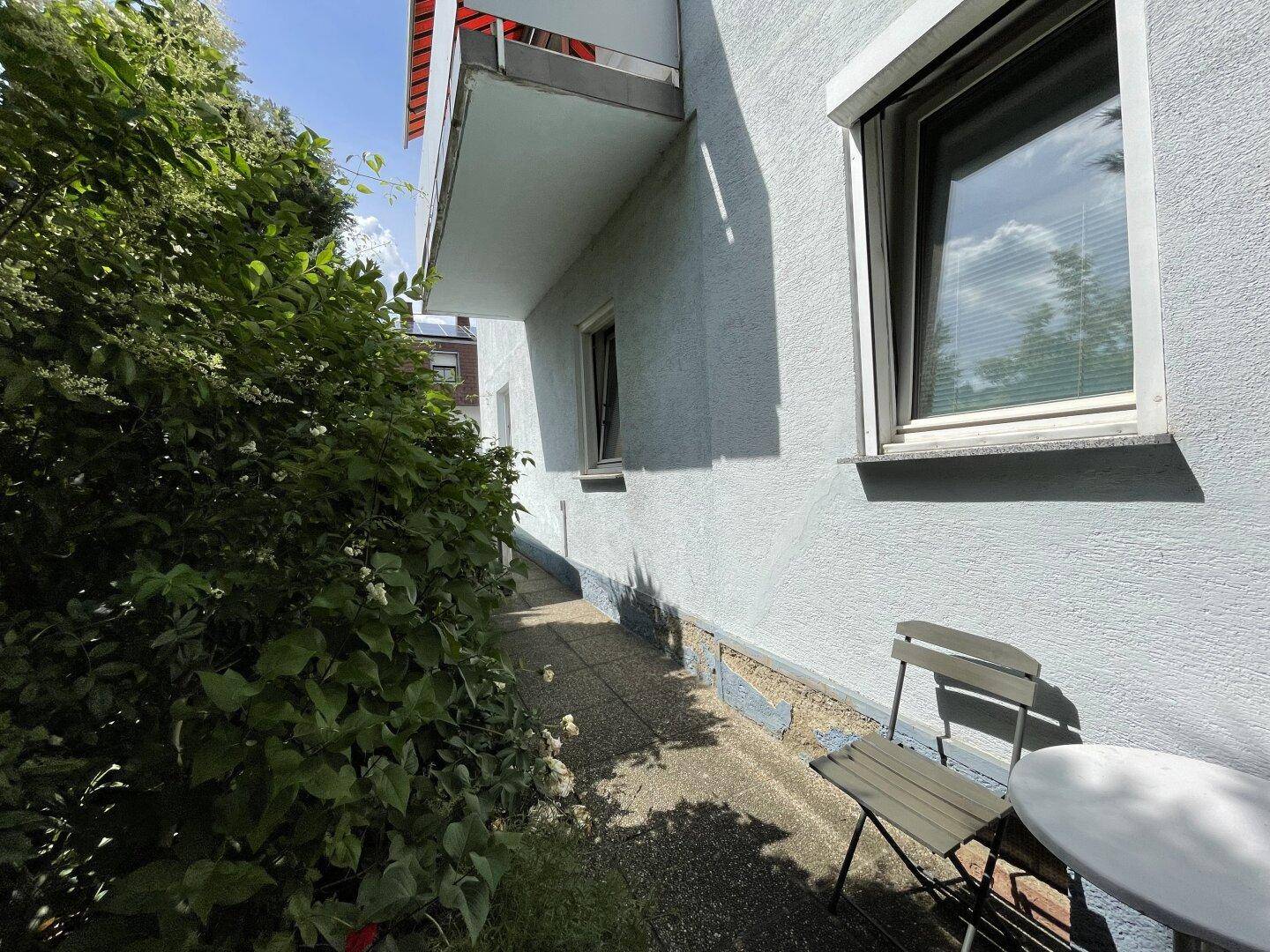 Schmaler Weg entlang der Hauswand mit üppigem Grün und einem kleinen Balkon darüber.
