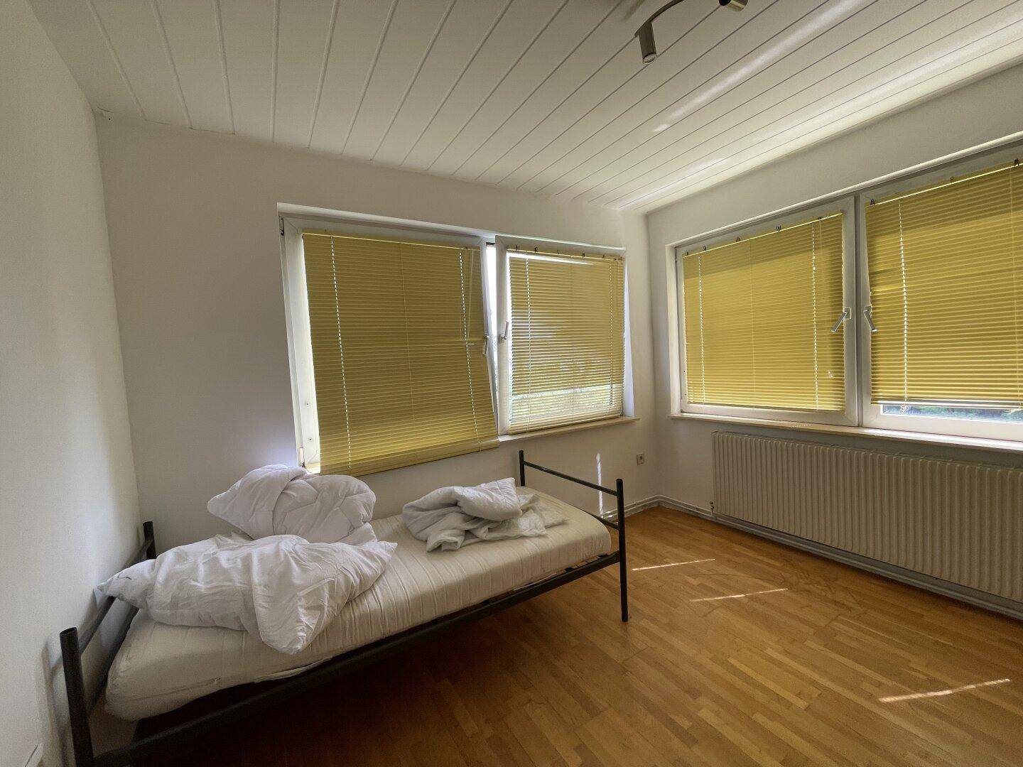 Helles Schlafzimmer mit Holzboden, Holzdecke und mehreren Fenstern mit Jalousien.