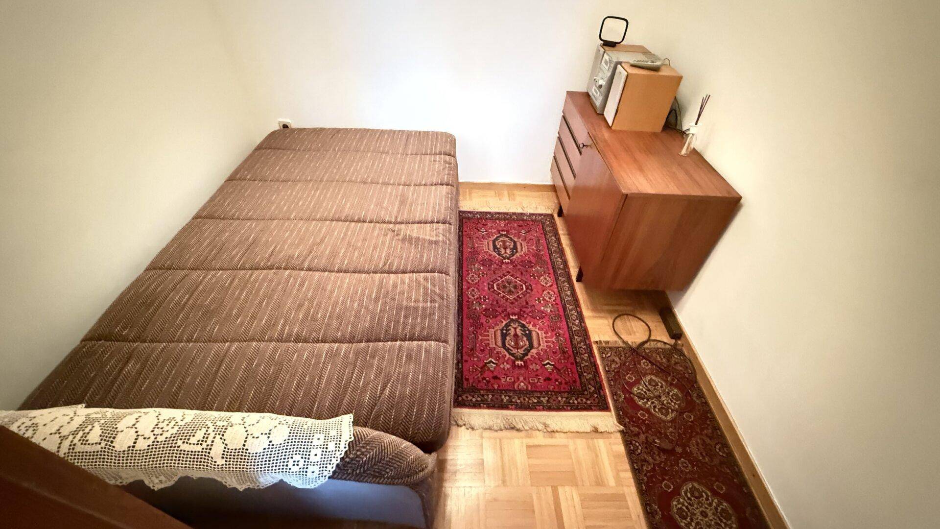 Kleines Schlafzimmer mit Einzelbett, Holzkommode und Teppich auf Parkettboden.