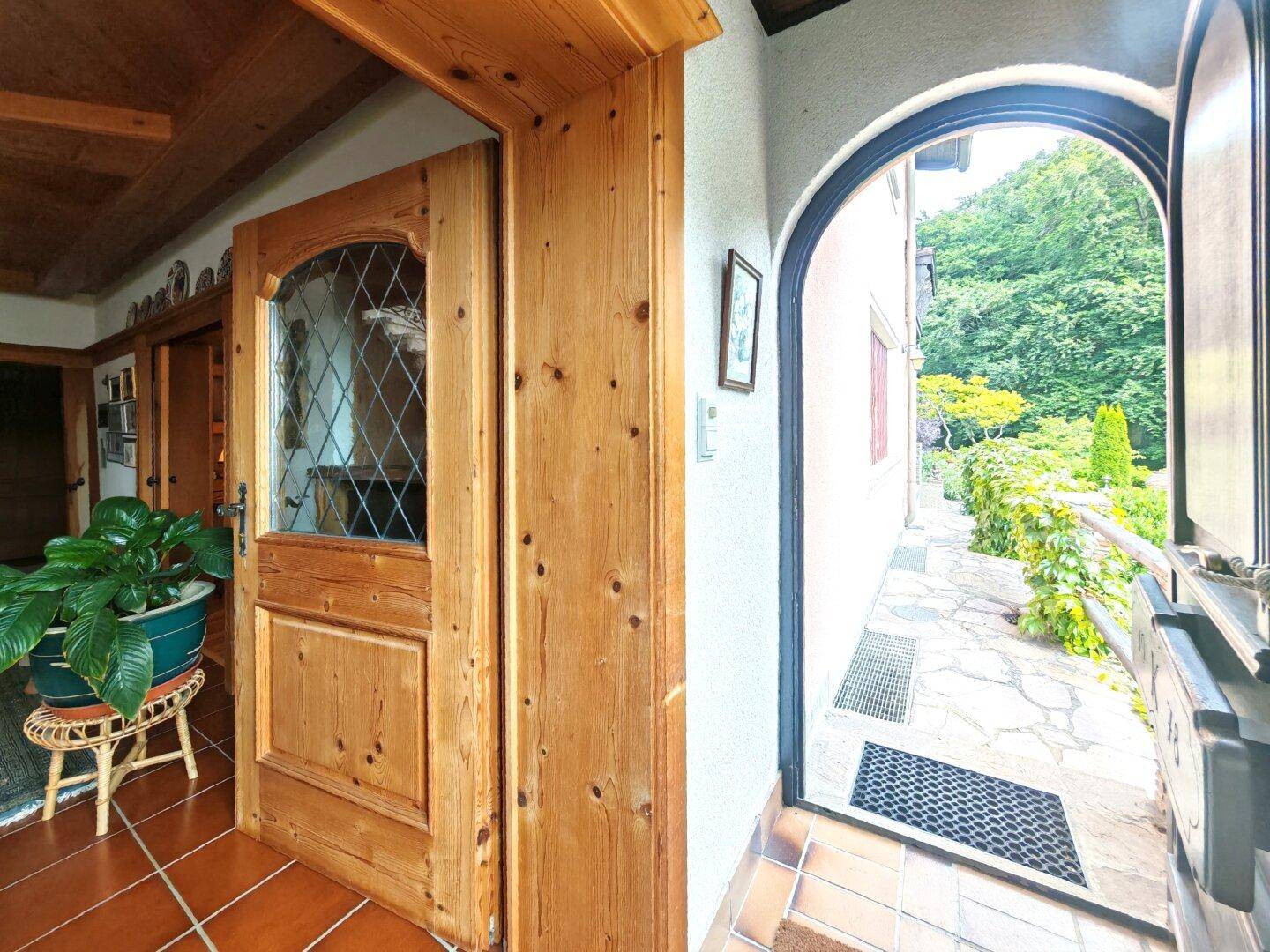 Heller Eingangsbereich mit Holztür, Fliesenboden und Blick nach draußen auf einen gepflasterten Weg.