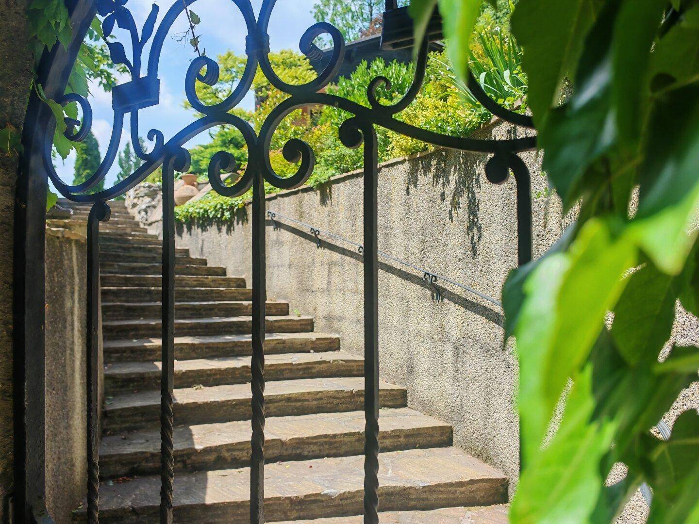 Detailansicht des schmiedeeisernen Tors und der Steintreppe, umgeben von üppigem Grün.