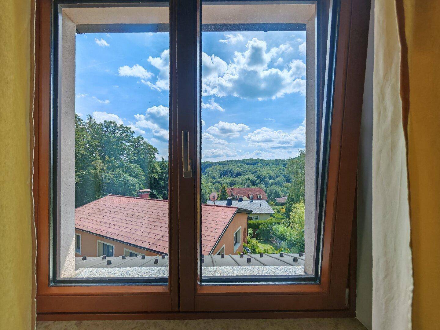 Ausblick aus einem Fenster auf Dächer, grüne Hügel und einen weiten blauen Himmel.