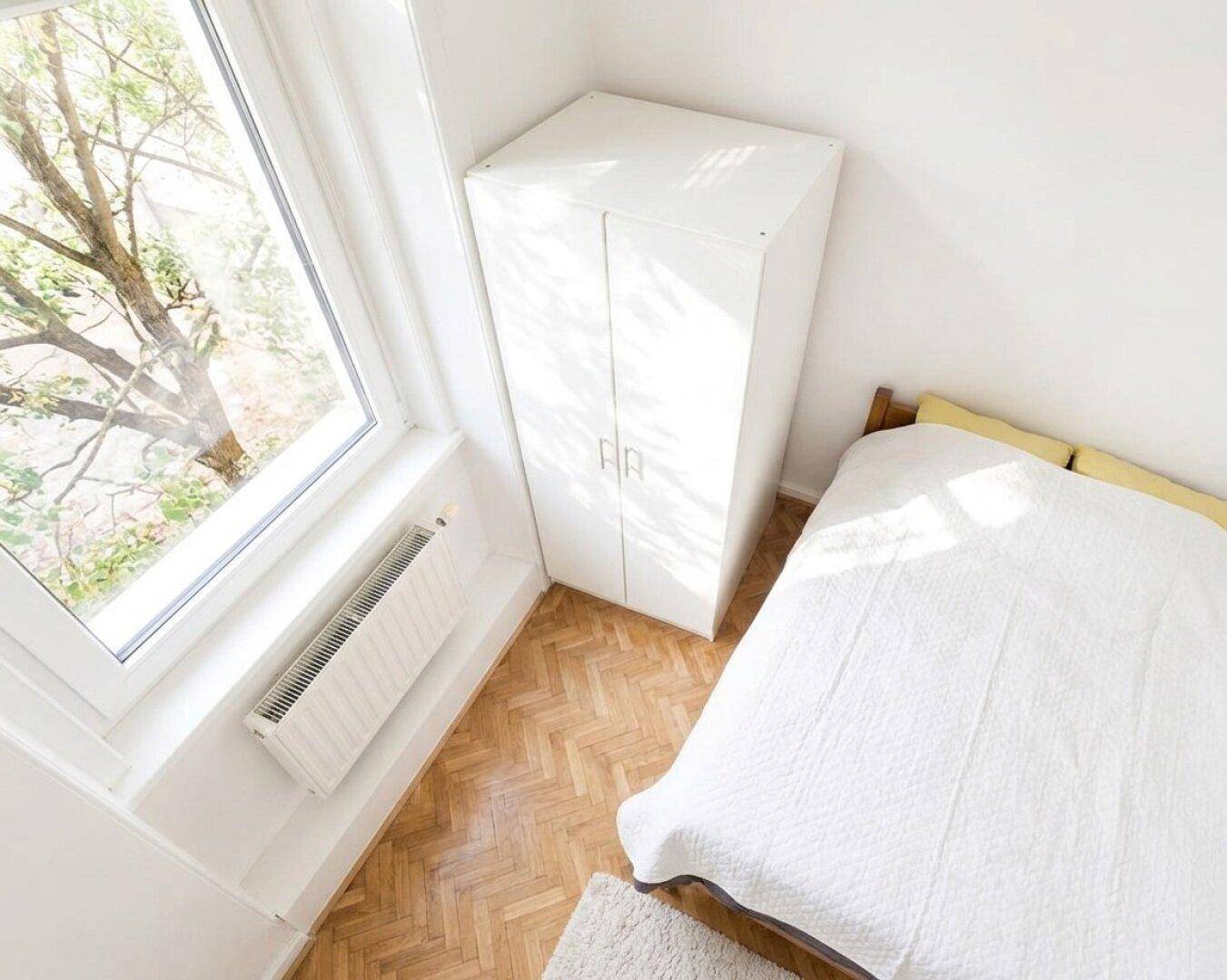 Helles Schlafzimmer mit Fischgrätparkett, weißem Kleiderschrank und Fenster mit Heizkörper.