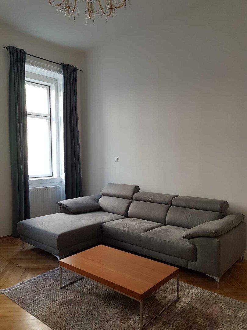 Modernes Ecksofa mit Holztisch und dunklen Vorhängen vor einem hohen Fenster.