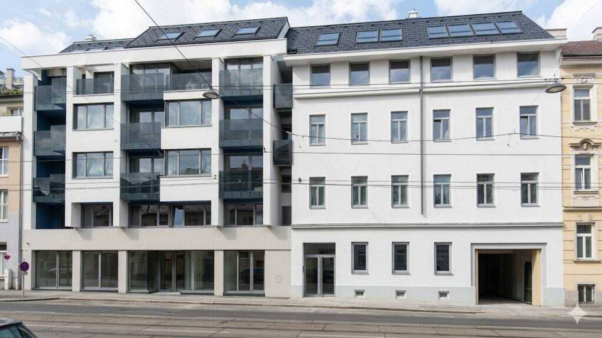 Moderne Gebäudefassade mit Balkonen und großen Fenstern neben einem älteren Wohnhaus an der Straße.