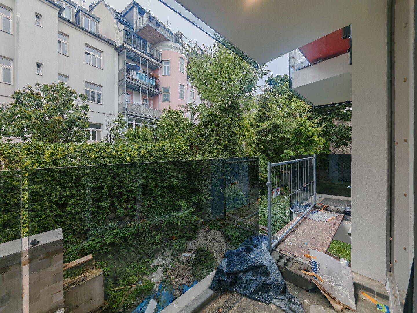 Blick von einem im Bau befindlichen Balkon auf grüne Hecken und umliegende Gebäude.