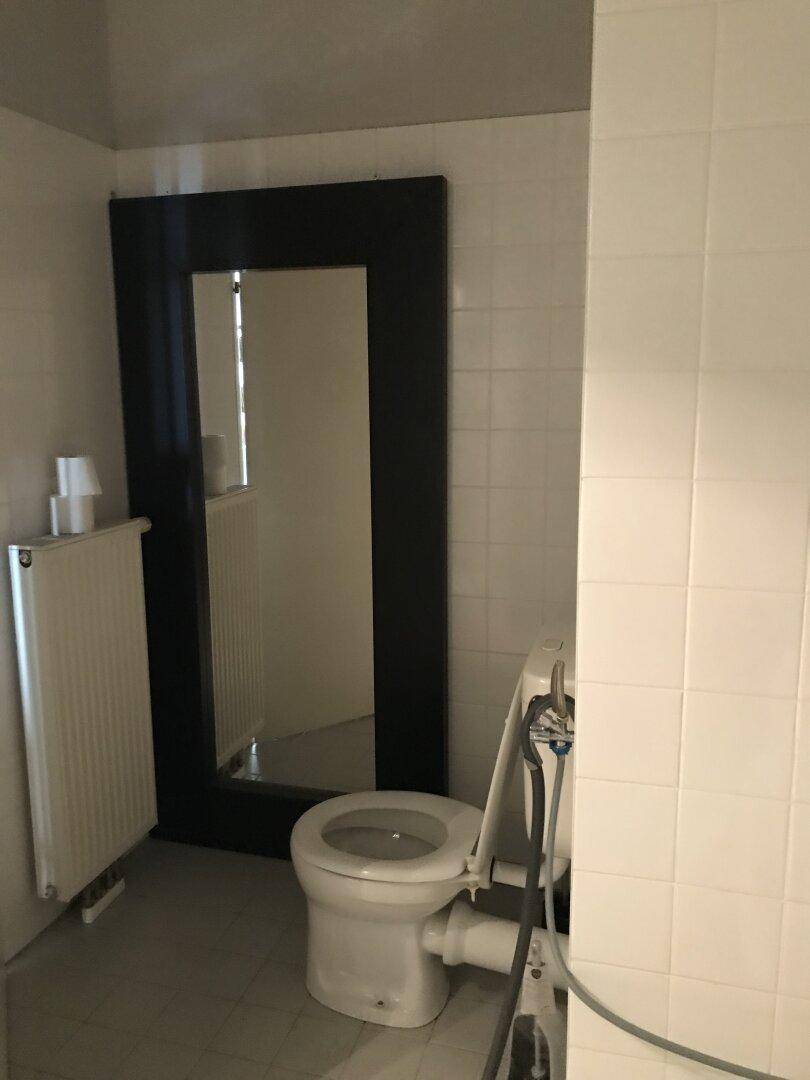 Toilette mit Heizkörper und großem Spiegel, in einem einfach gefliesten Raum.