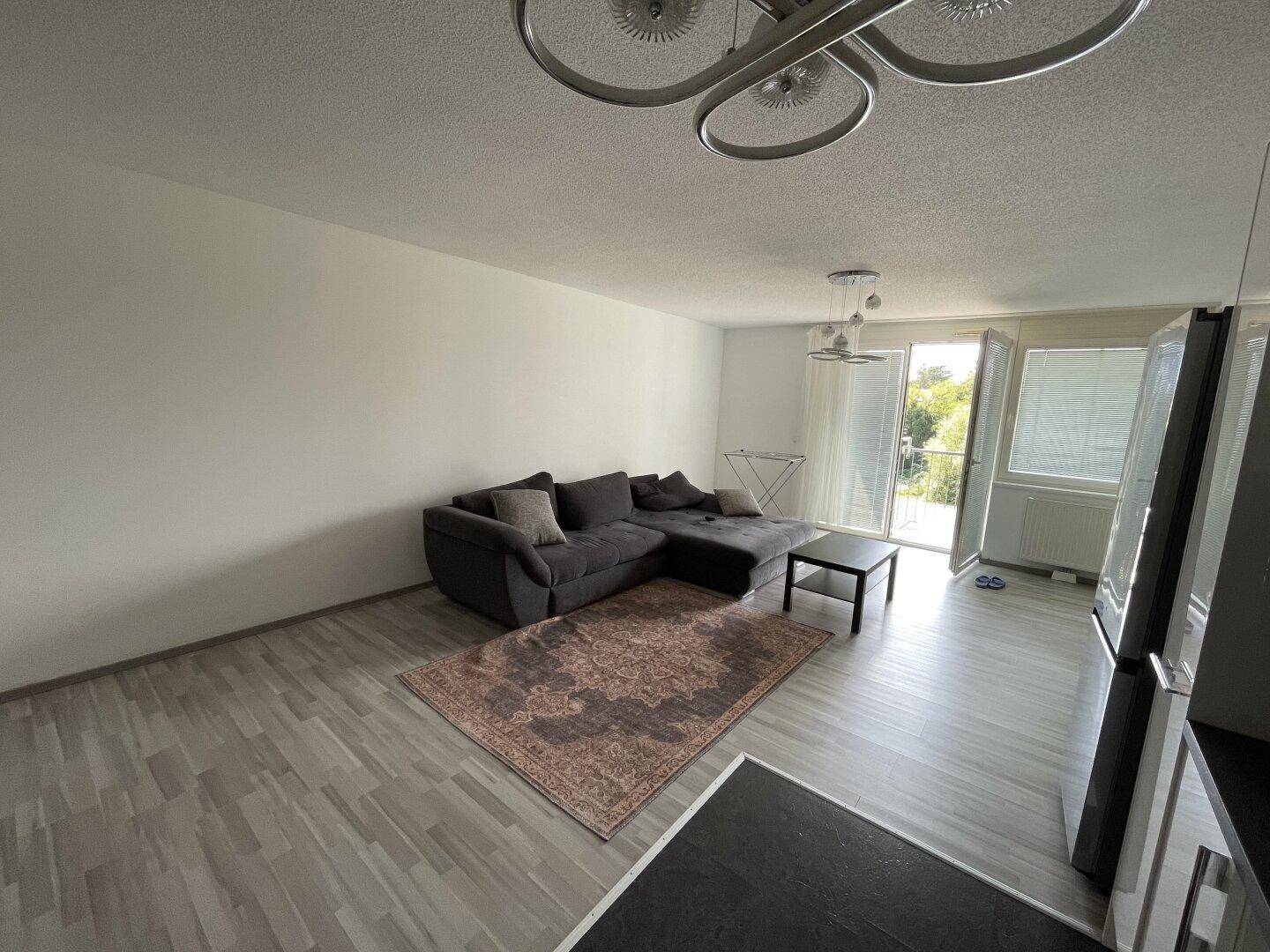 Heller Wohnbereich mit grauem Ecksofa, Teppich und Zugang zum Balkon.