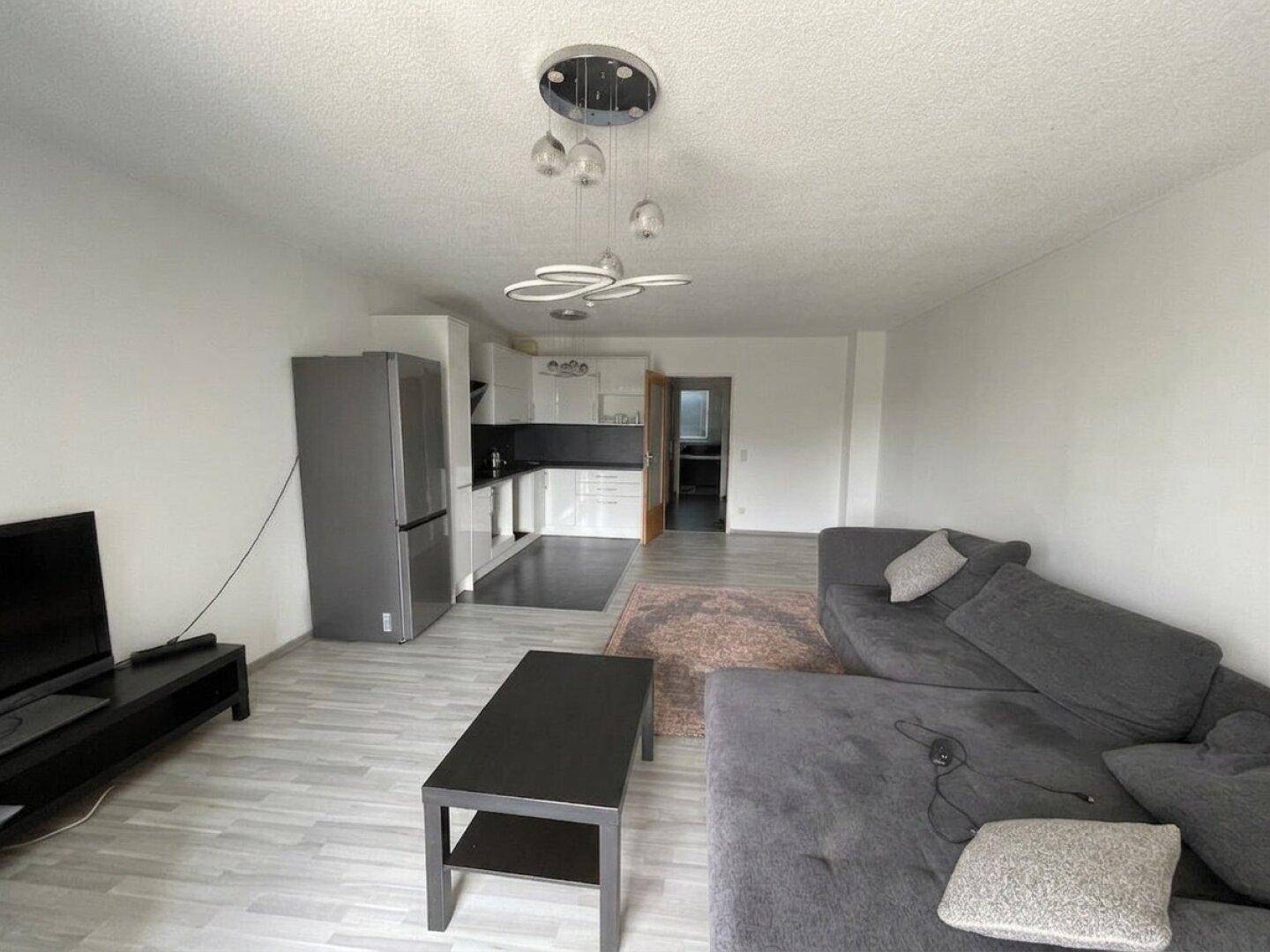 Offener Wohnbereich mit integrierter Küche, grauem Sofa und hellem Laminatboden.