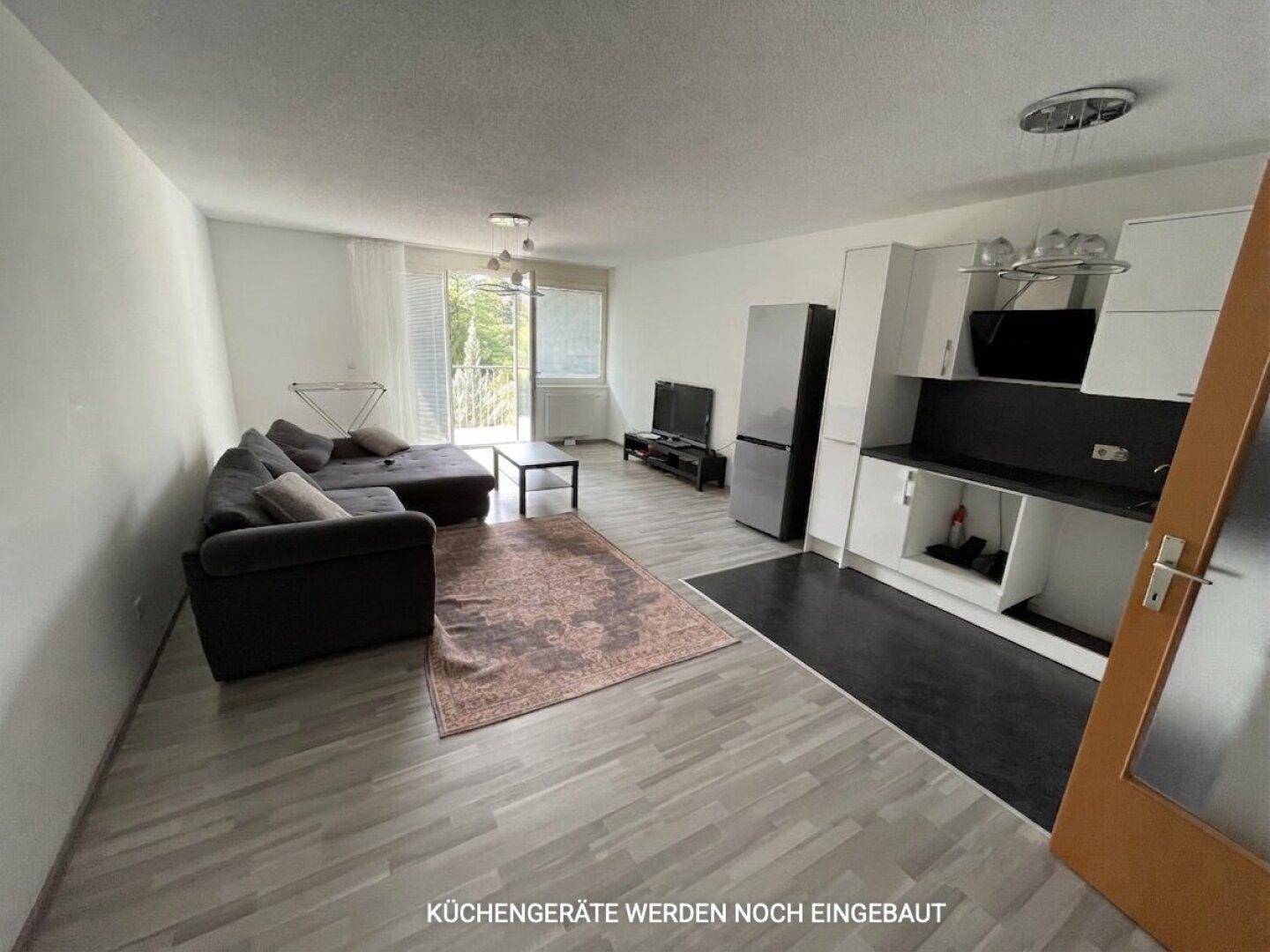 Offener Wohn- und Küchenbereich mit grauem Sofa; Küchengeräte werden noch eingebaut.