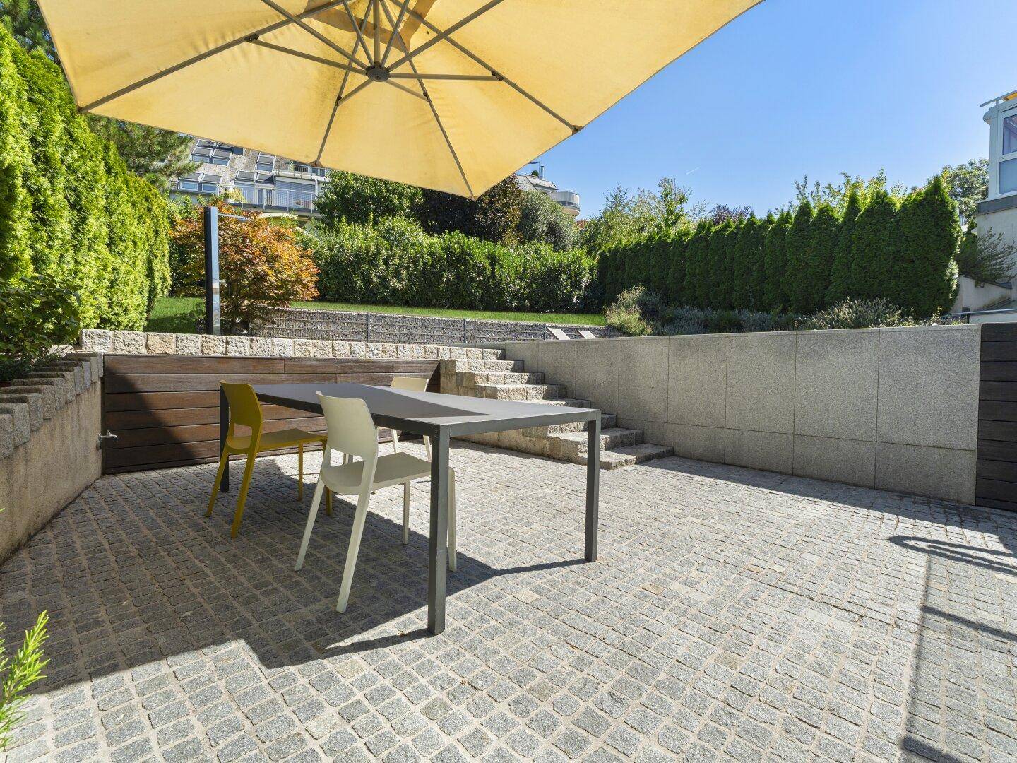 Gepflasterte Terrasse mit Esstisch und Stühlen unter einem großen Sonnenschirm, umgeben von Mauern.