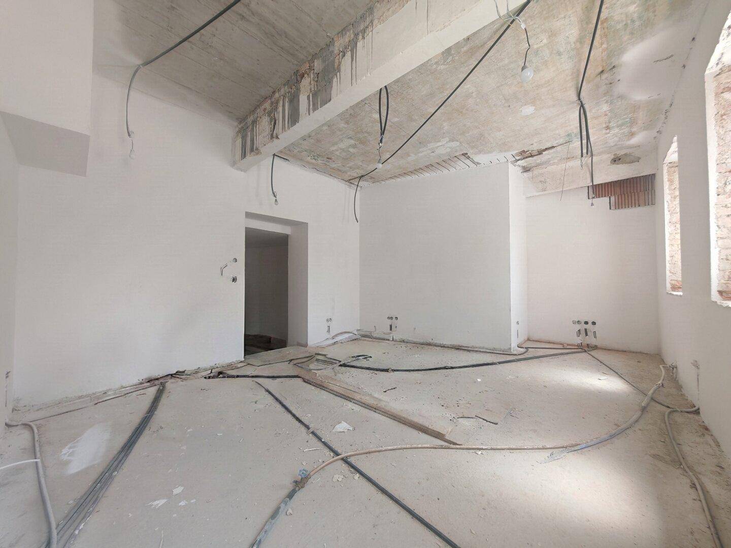 Rohbau eines Raumes mit sichtbaren Kabeln an der Decke und unfertigen Wänden und Boden.